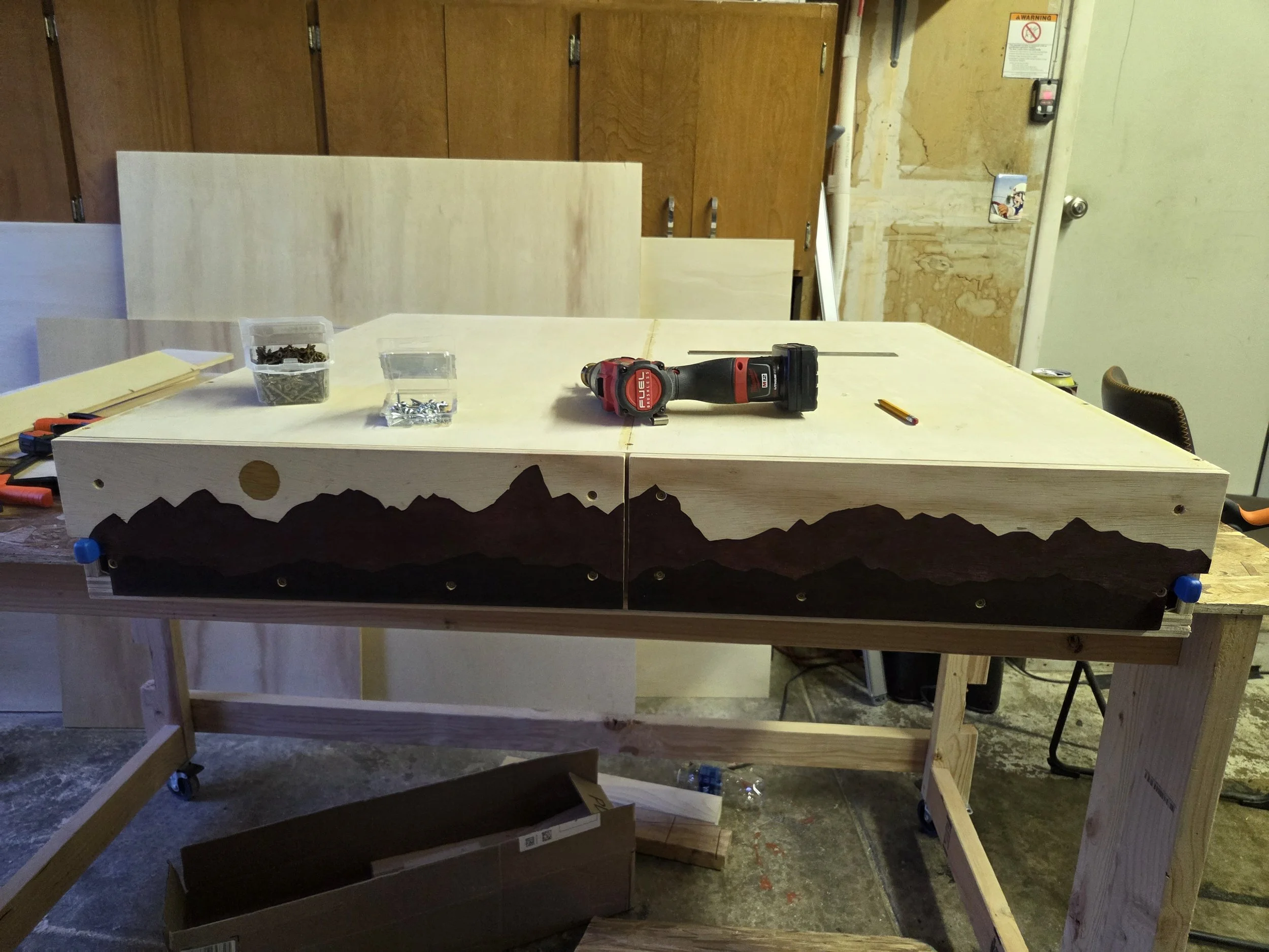 A work-in-progress wooden table with a mountain silhouette design on the side, attached to a sturdy wooden frame. Tools, screws, and a cordless drill are placed on the table, indicating ongoing carpentry or woodworking project in a workshop with exposed wall patches, cabinets, and safety signs.
