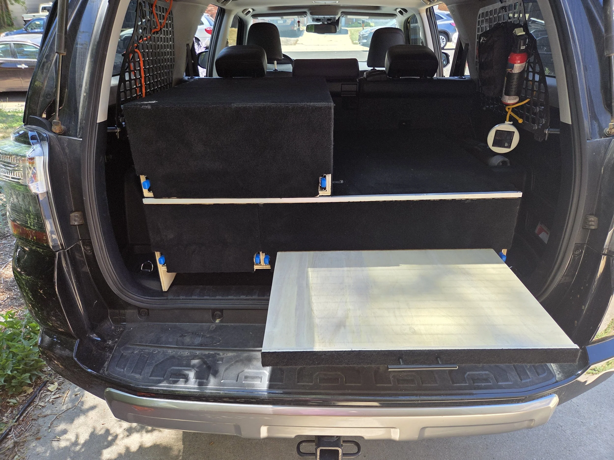 The back of a black SUV with its liftgate open, revealing a cargo area. The cargo space is equipped with a gray metal platform and a small black carpeted partition. A fire extinguisher is mounted on the right side, attached to a black grid panel. Several parked cars are visible in the background.