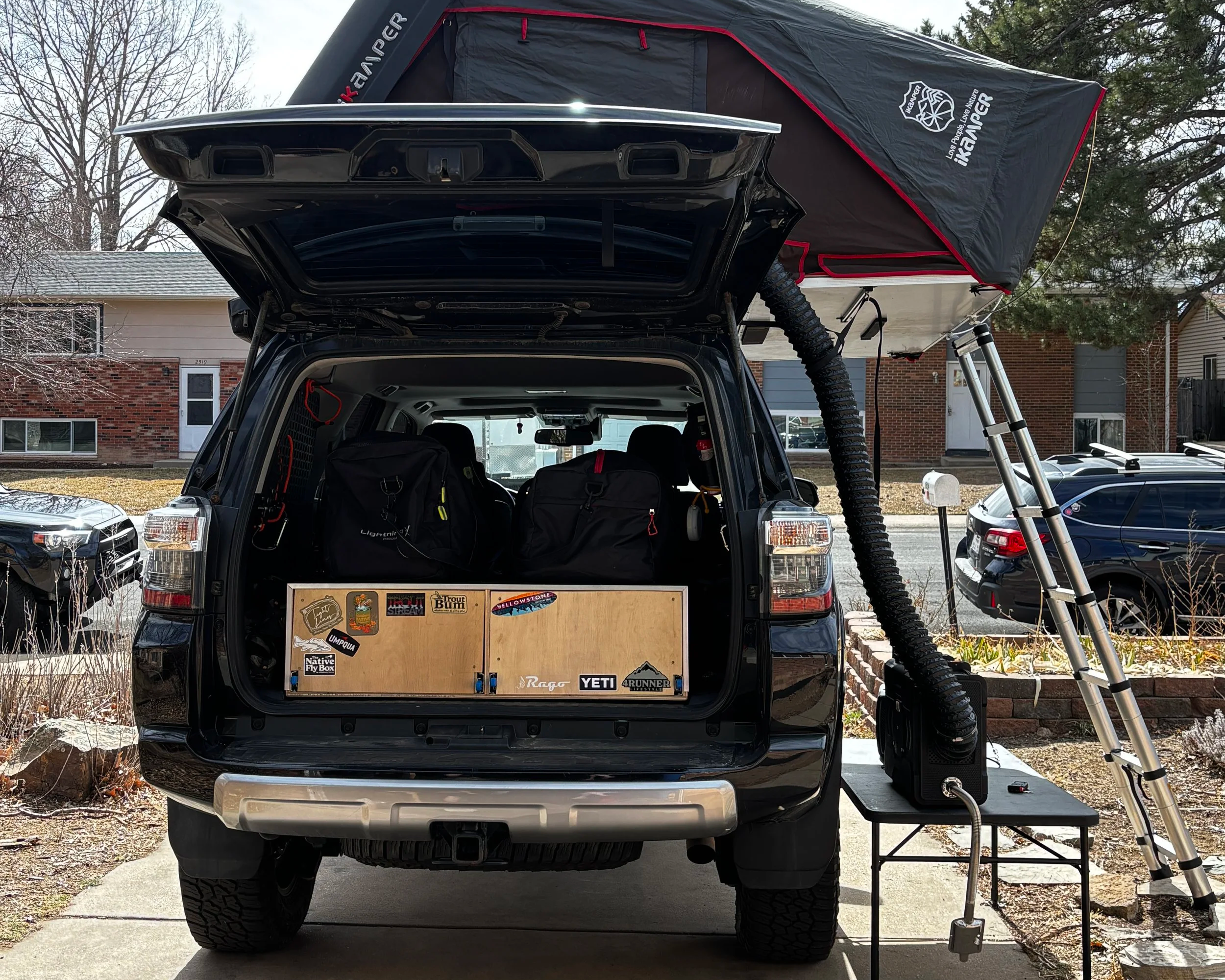 Back of a black SUV with open tailgate showing two backpacks inside, attached to a rooftop tent with a metal frame and ladder, parked in a residential neighborhood with houses and cars visible.