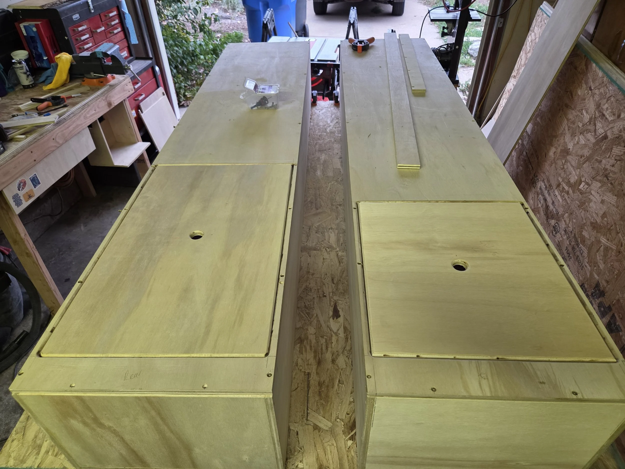 Wooden workbenches with yellow painted surface in a garage workshop, tools and materials on the benches, open garage door with outdoor view.