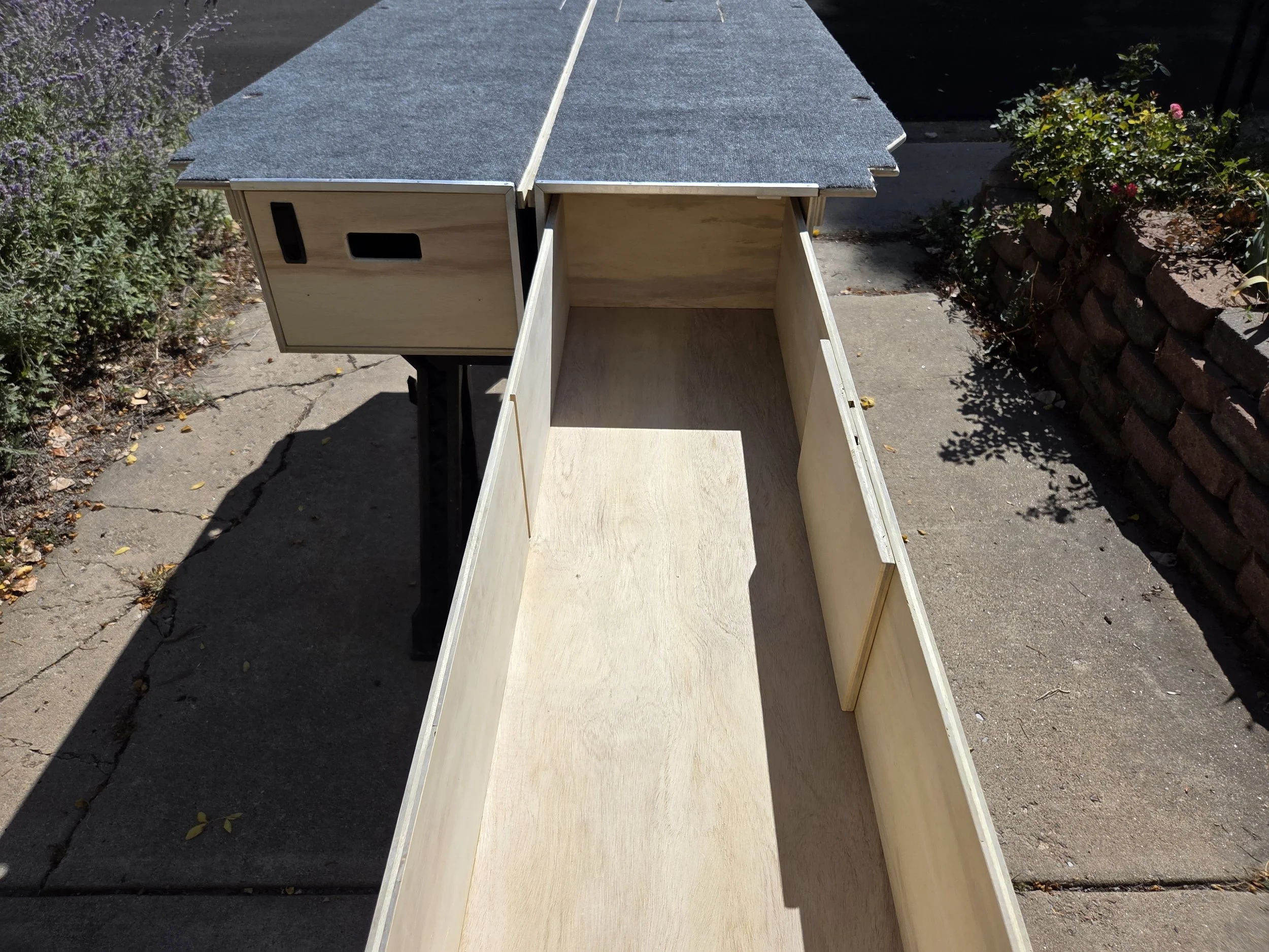 Unfinished wooden structure, possibly a small cabinet or box, on a concrete sidewalk outdoors with plants and flowers on either side.