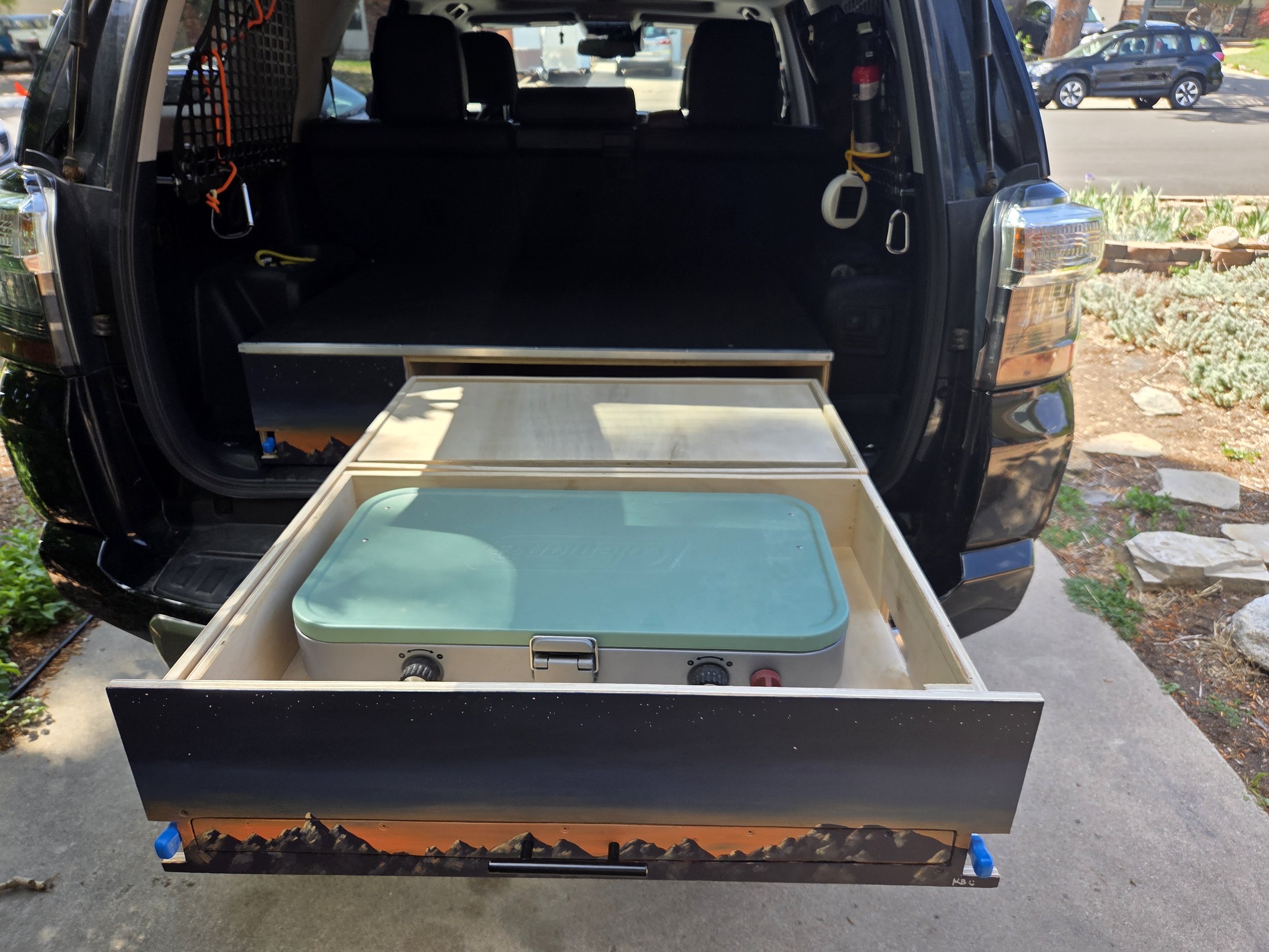 Black SUV with open rear hatch showing interior prep for camping or travel with a wooden drawer and cooler in the foreground.
