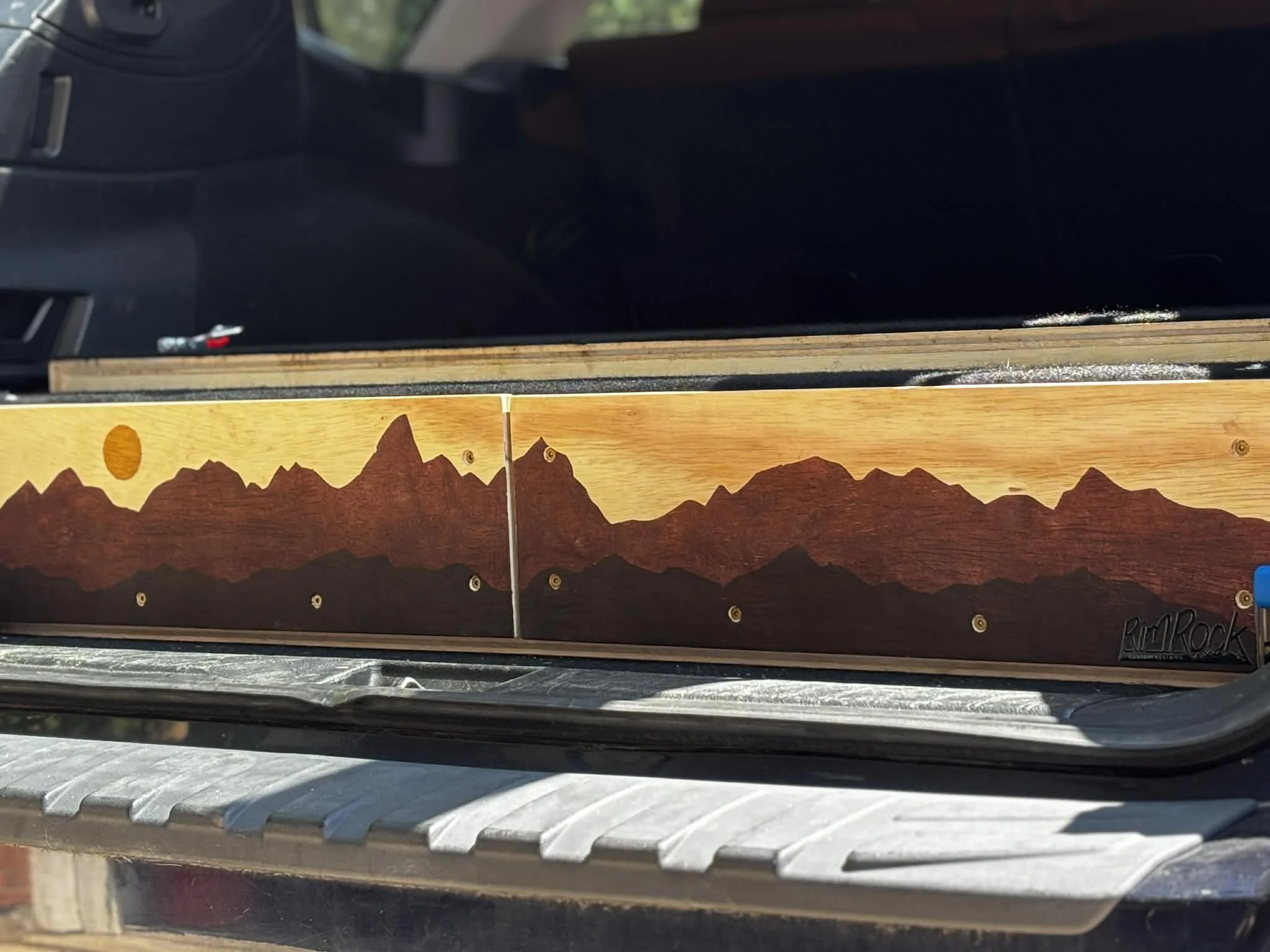 Close-up of a wooden sign with painted mountains and a bright yellow sun, placed on a metal surface with a black object and a window in the background.
