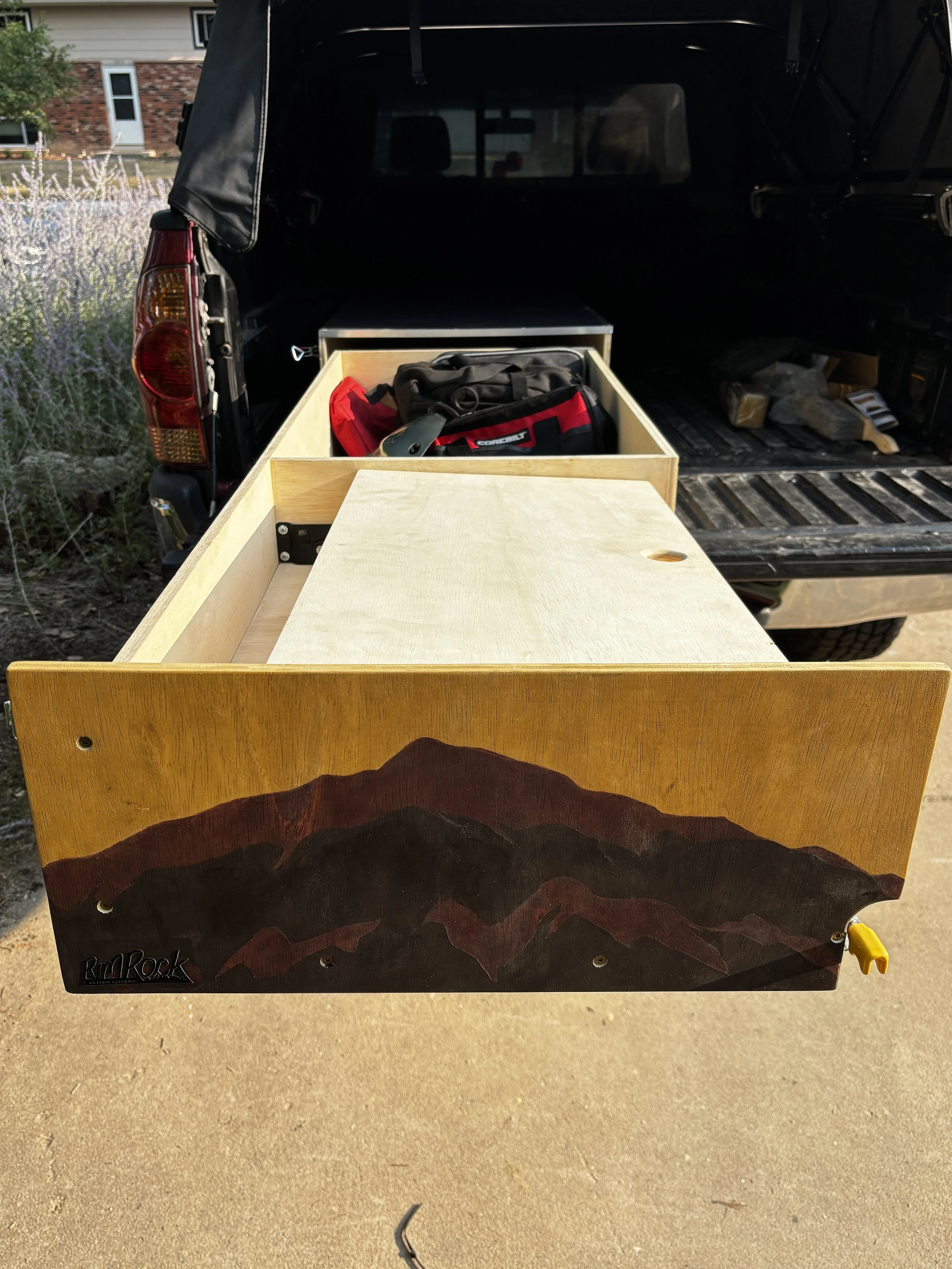 A black pickup truck with an open bed containing a partially assembled wooden cabinet, a red and black tool bag, and some miscellaneous items. The scene is outdoors in front of a house with a brick and siding exterior, and a garden with plants on the left.