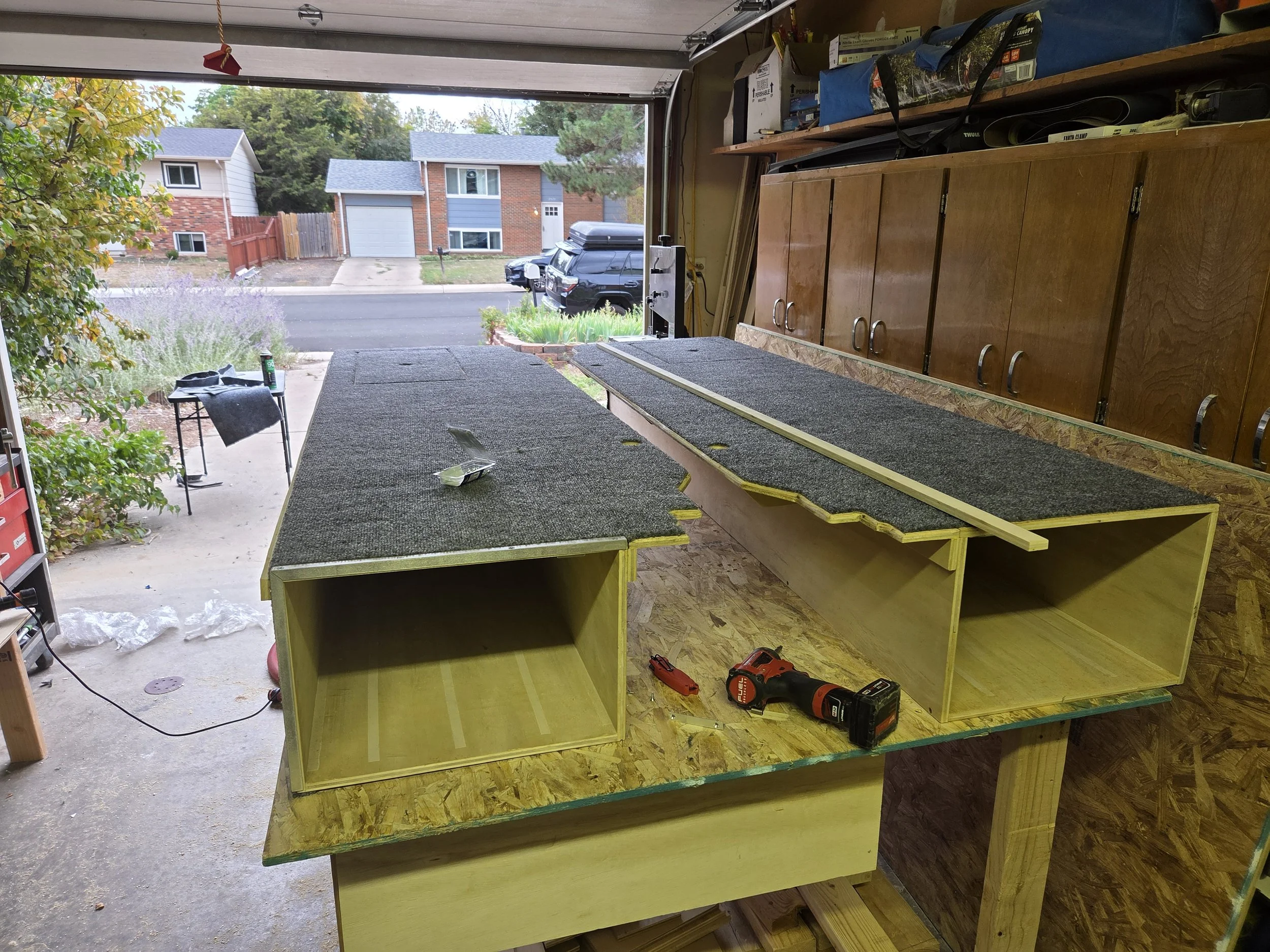 View of a garage interior with an unfinished wooden workbench, tools, and supplies. The garage door is open, revealing a suburban street with houses, cars, and greenery outside.