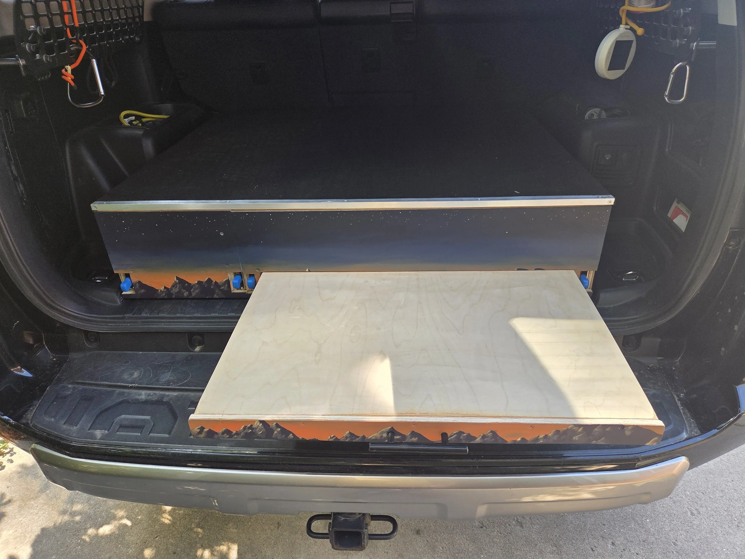 Inside the trunk of a car, there is a black wooden platform with a mountain landscape design at the bottom. The trunk contains some tools and equipment, including a white device hanging on the right side and some wires in the back. The car has a black interior with hook attachments and a car keyring on the left side.
