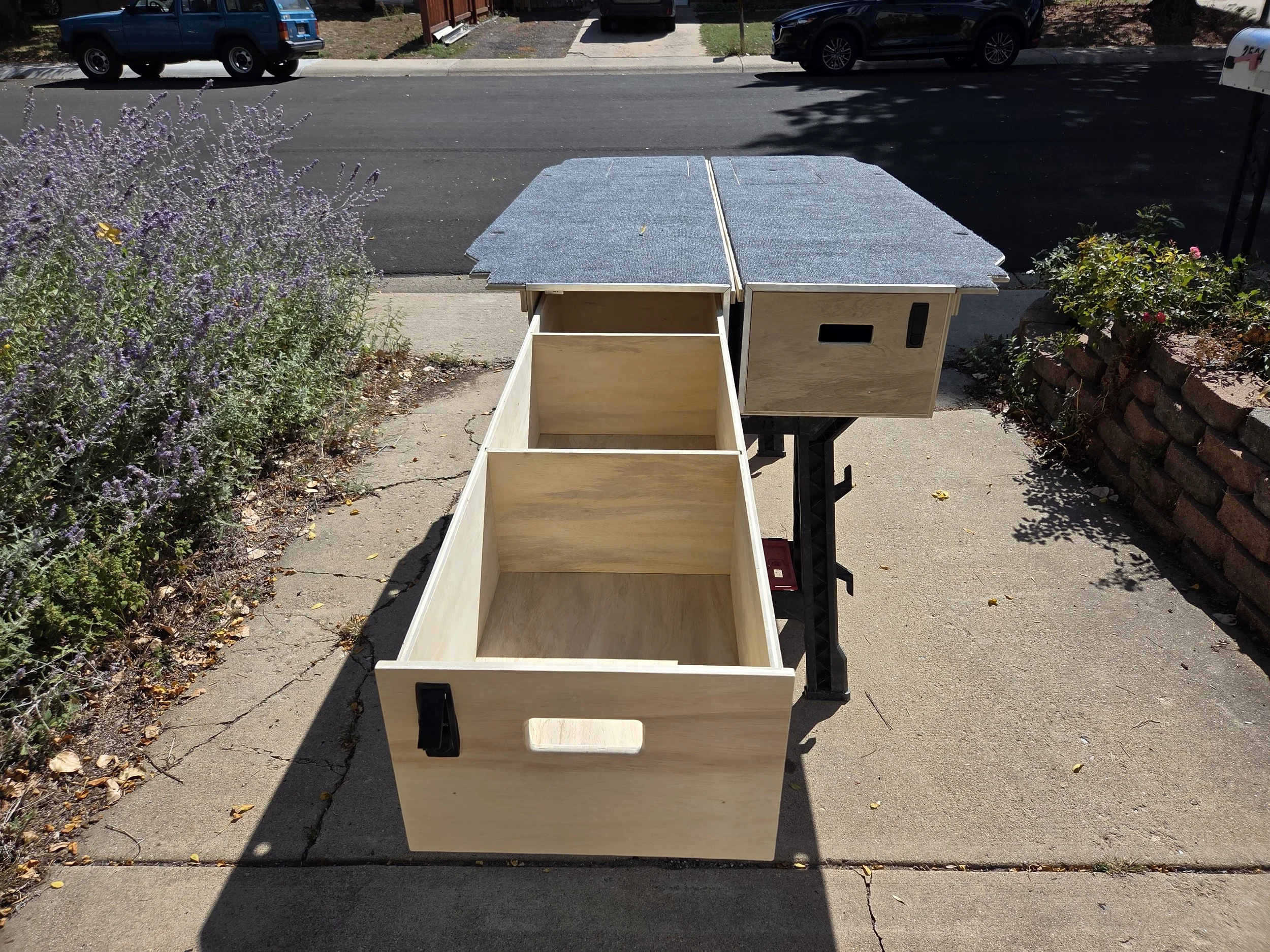 Unfinished wooden cabinet on a foldable table on a sidewalk, with cars parked and a mailbox in the background.