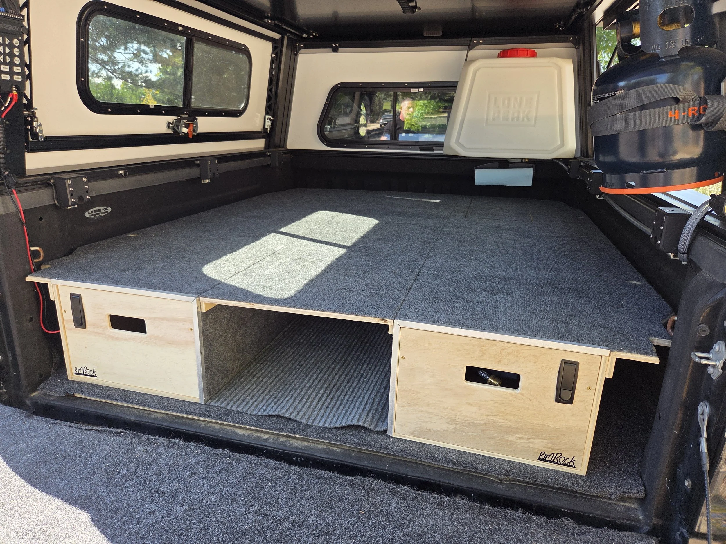 The interior of an off-road vehicle or camper, featuring a built-in wooden storage cabinet, a white water container, and various equipment, with windows showing a sunny outdoor scene.