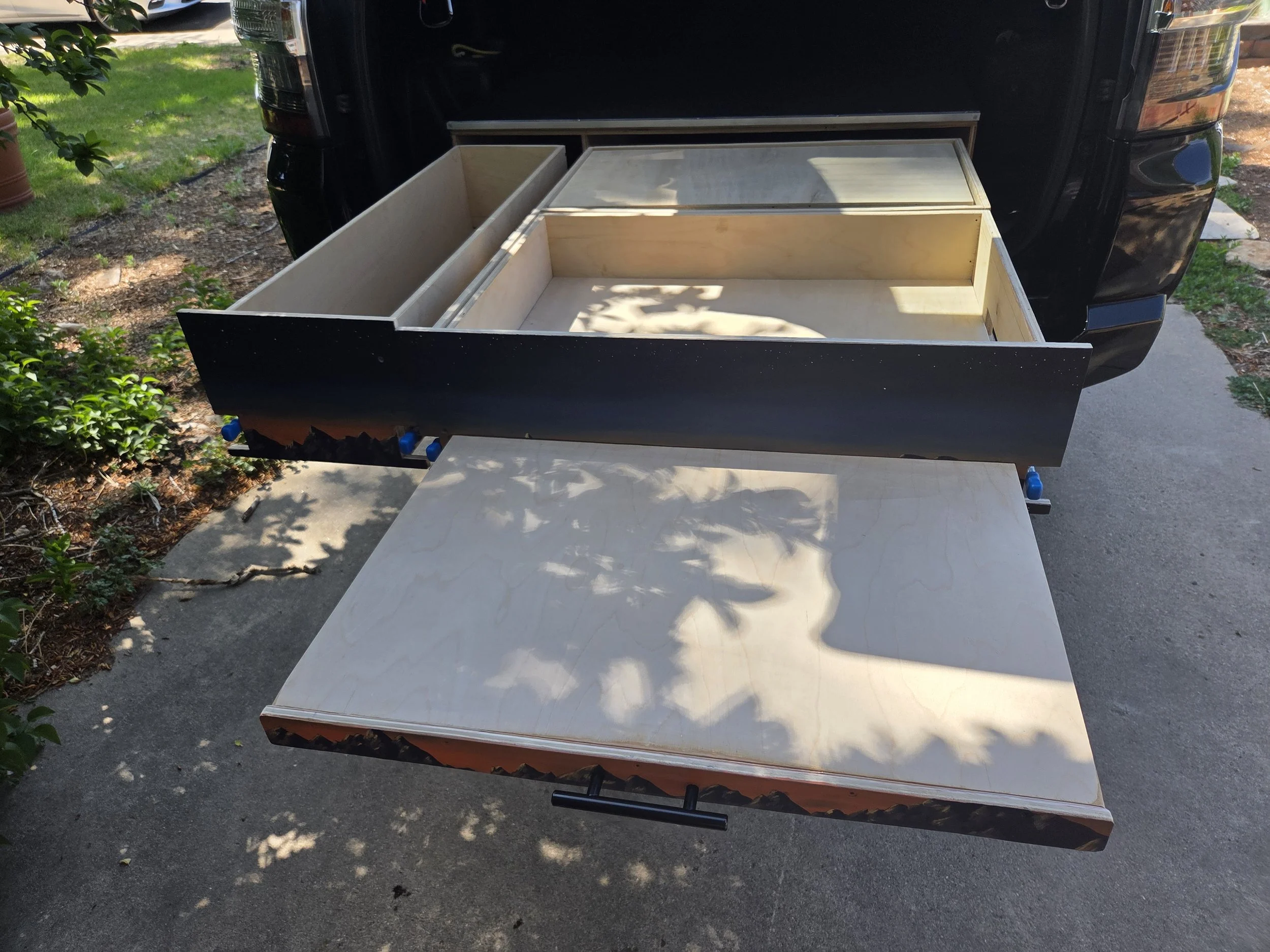 Wooden furniture piece, possibly a drawer or cabinet, placed outdoors on a table, with a black truck in the background and greenery surrounding the area.