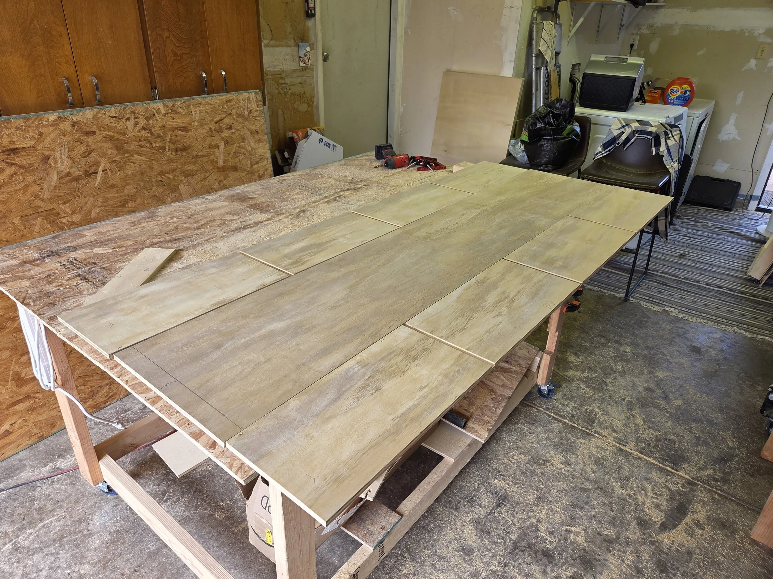 Workshop table with woodworking tools, sheets of wood, and construction materials in an unfinished room.