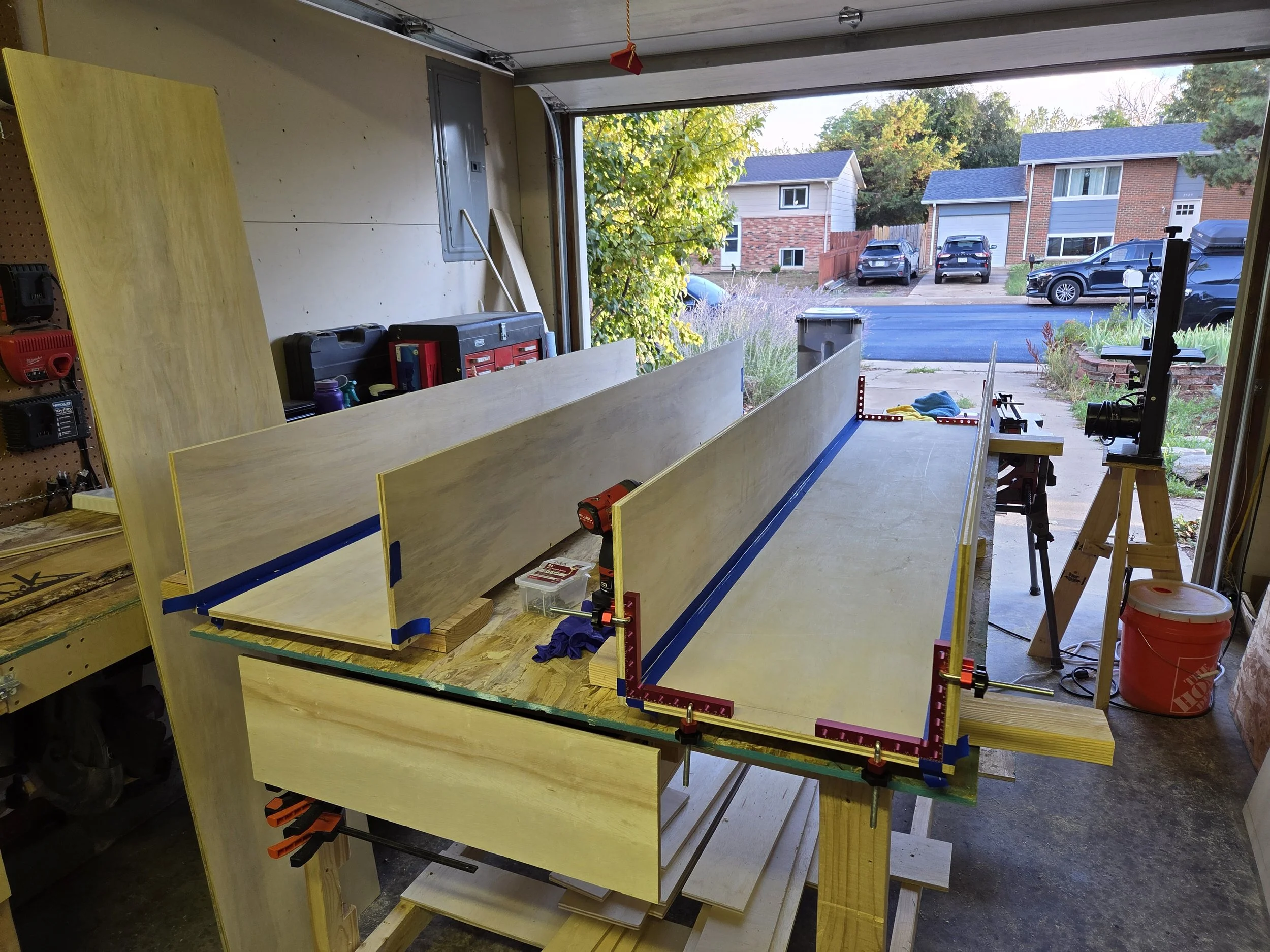 Inside a garage with woodworking tools and materials, showing partially assembled wooden panels, clamps, and a view of a residential street outside.