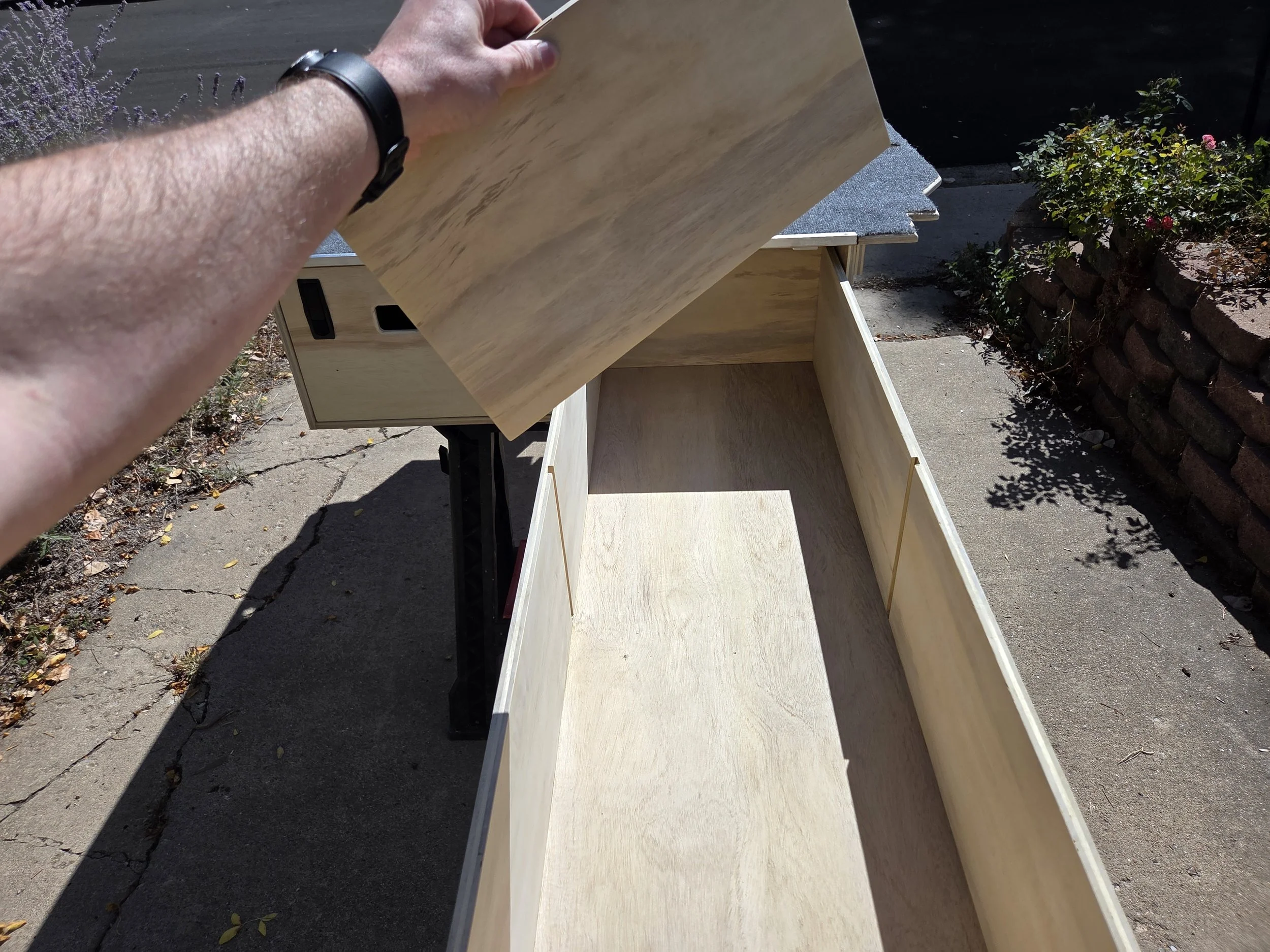 A person's hand holding a sheet of wood above a partially assembled wooden cabinet outdoors on a sunny day. The background includes bushes, a stone wall, and pavement.