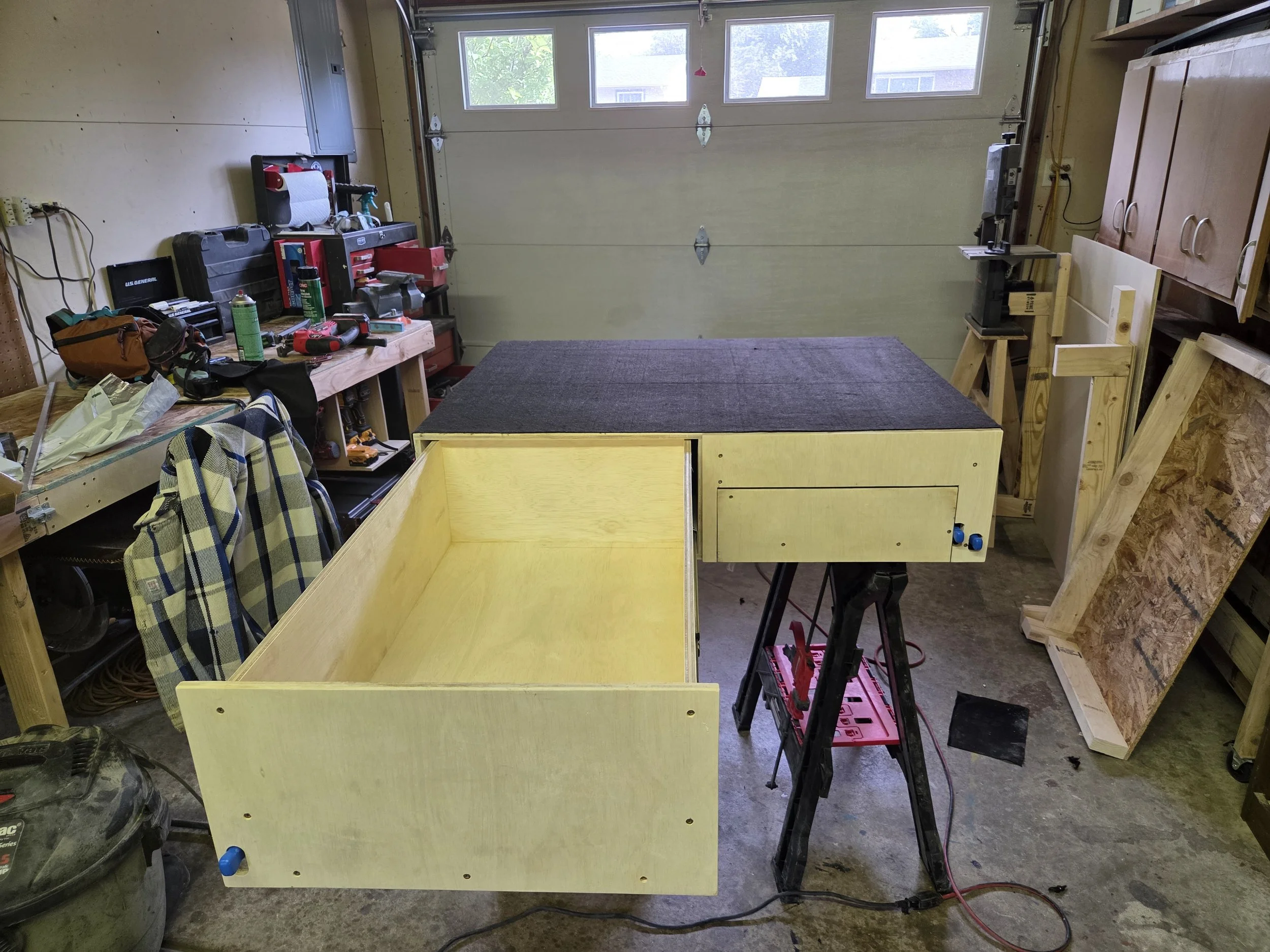 Garage workshop with tools and wooden furniture under construction. Workbench on left, saw on right, large garage door in background, and wooden parts leaning on shelf.