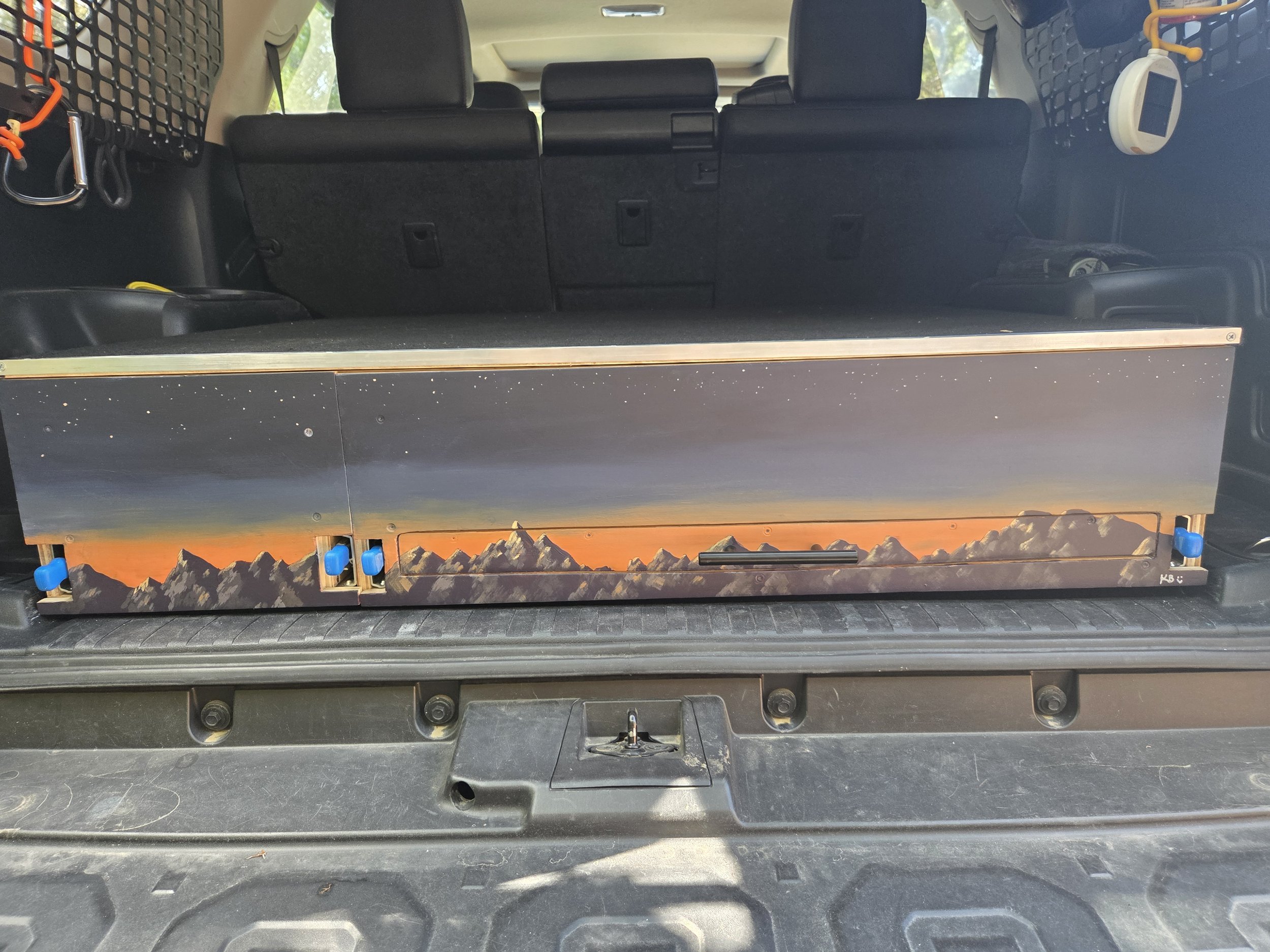 The back of a vehicle with a cargo area. The cargo area has a painted mountain landscape scene at the bottom. The interior is organized with mesh storage and tools.