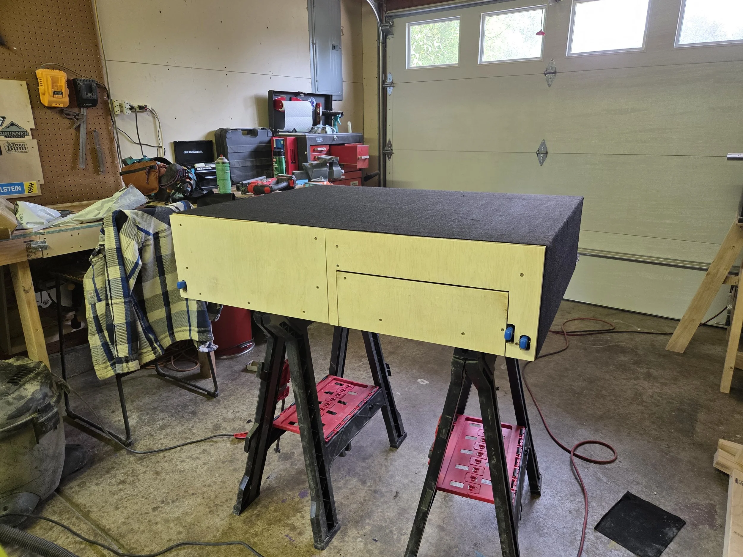 A garage workshop with tools, a black workbench, a yellow wooden worktable on sawhorses, and a partially open garage door with windows.