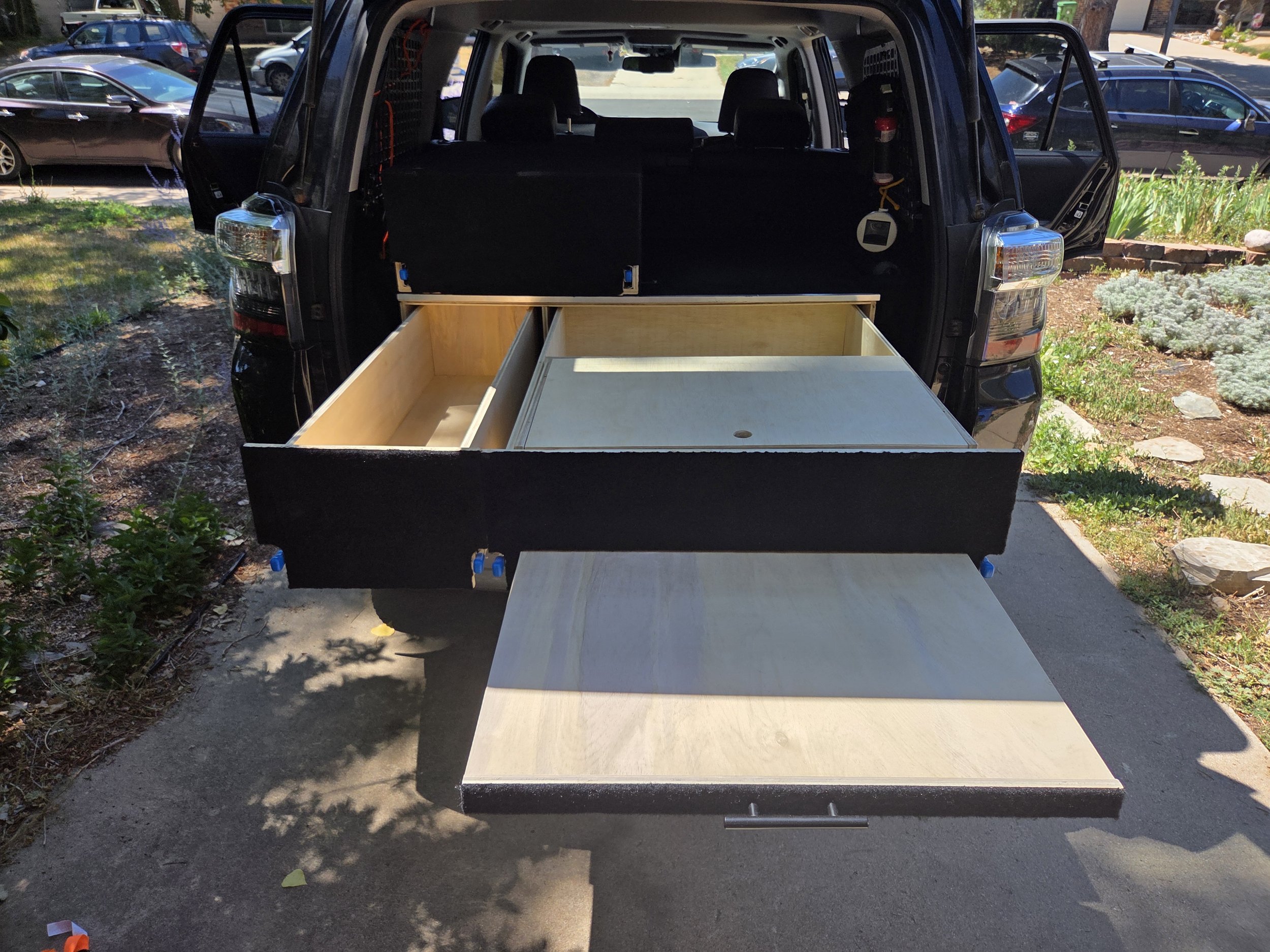 Black SUV with an open rear hatch, showing an unfinished wooden drawer system in the cargo area, parked on a driveway with other cars and a garden with plants and flowers nearby.