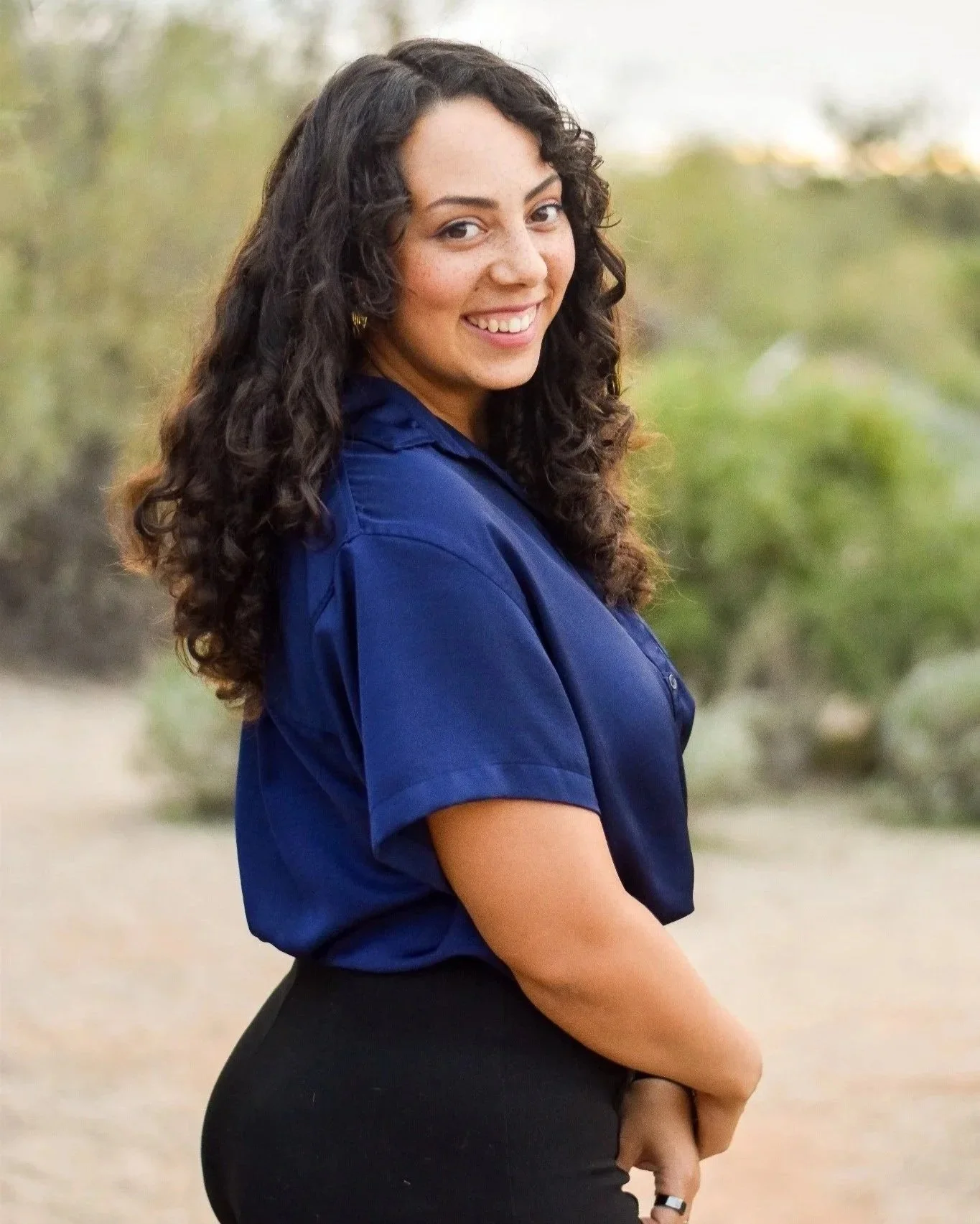 A woman with long curly dark hair smiling outdoors, wearing a blue shirt and black pants, with blurred greenery in the background.