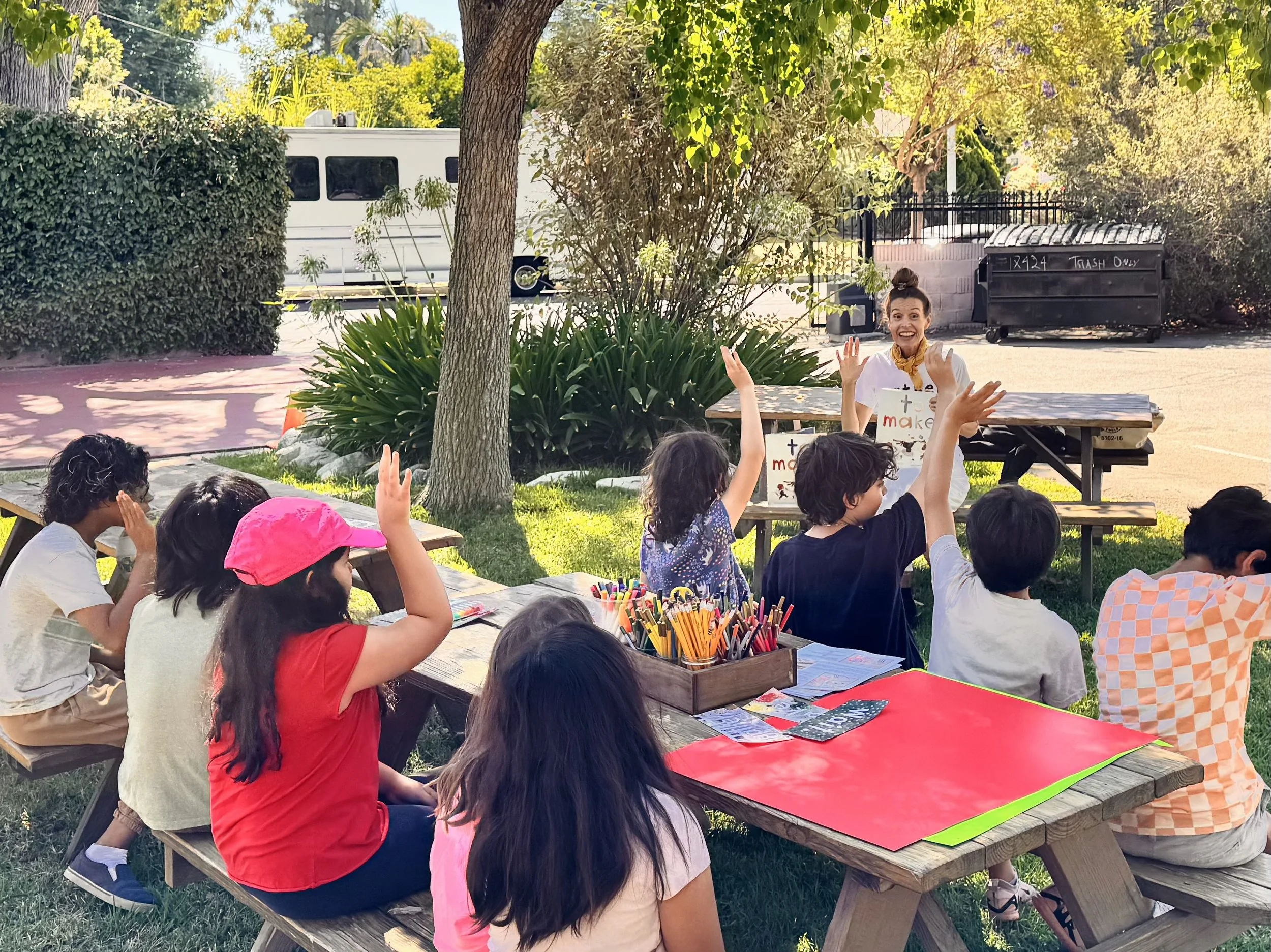 A woman standing outdoors, smiling and speaking to a group of children seated at picnic tables, raising her hands, with trees, bushes, and a parked van in the background.