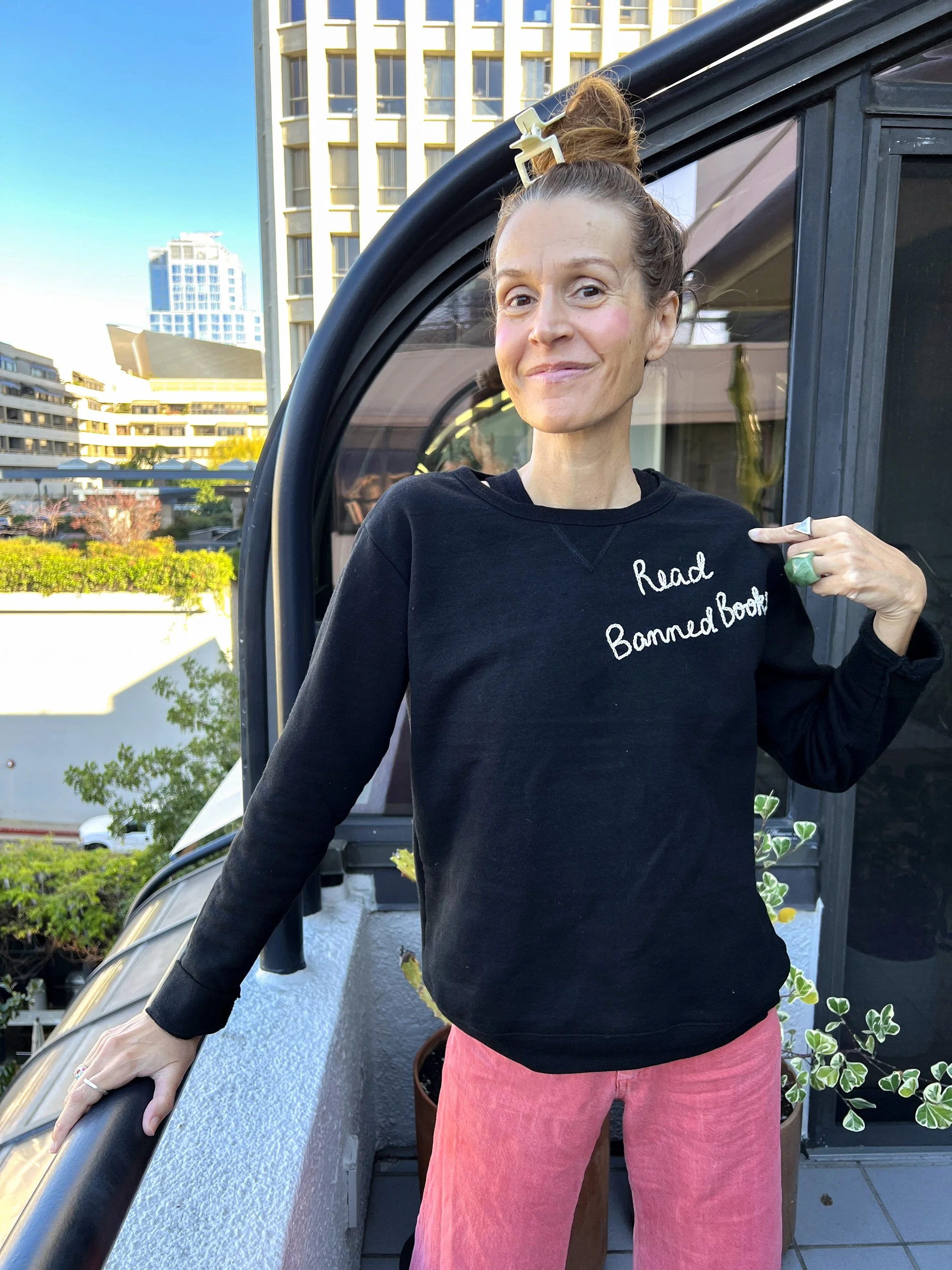 A woman standing outdoors, leaning against a black railing, wearing a black sweatshirt with white text, pink pants, and a large ring on her finger. Her hair is tied up with a beige hair clip, and she has a slight smile. The background features residential buildings, trees, and a clear blue sky.