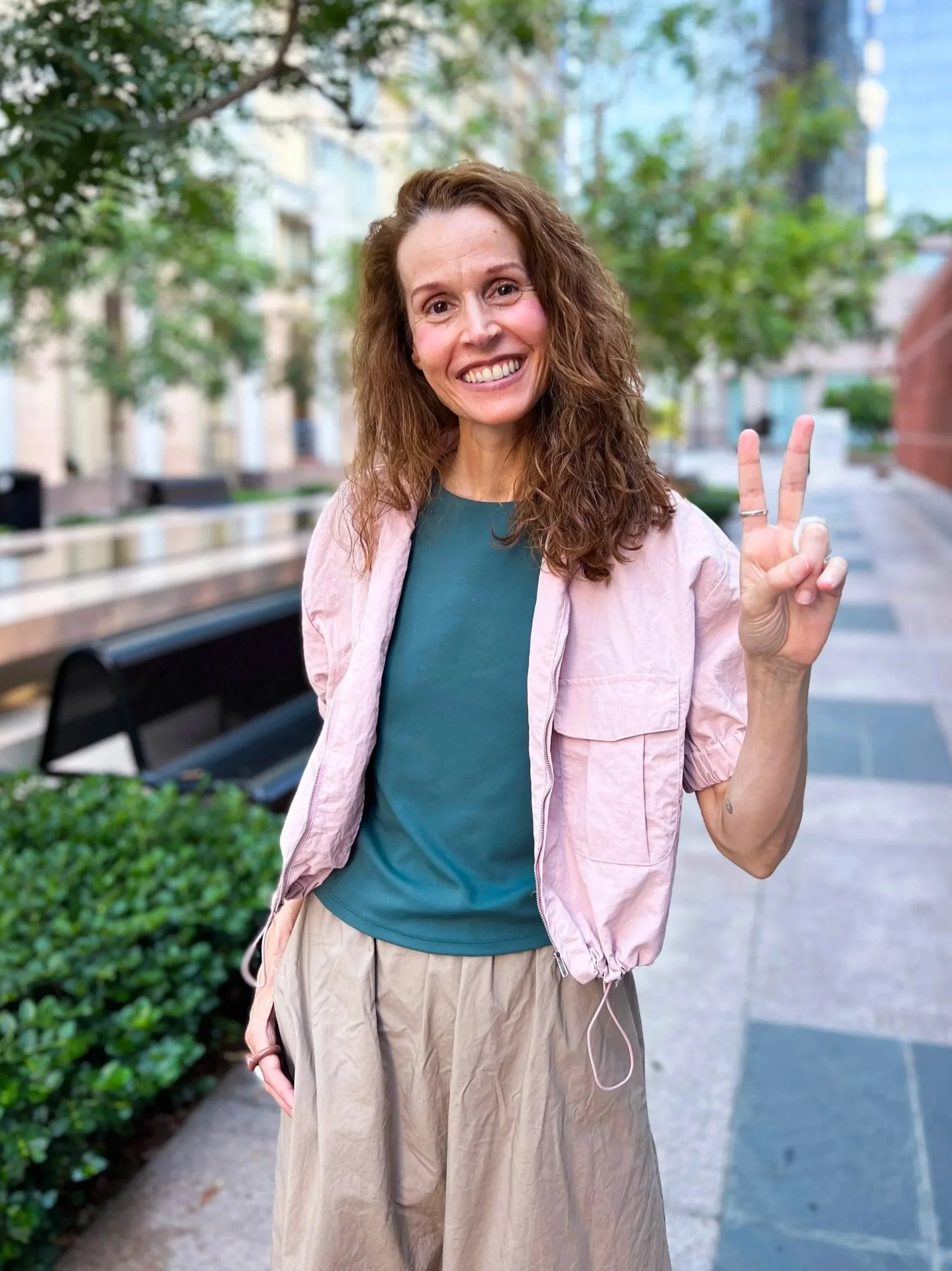 A smiling woman with curly red hair making a peace sign with her right hand outdoors in an urban setting with trees and buildings in the background.