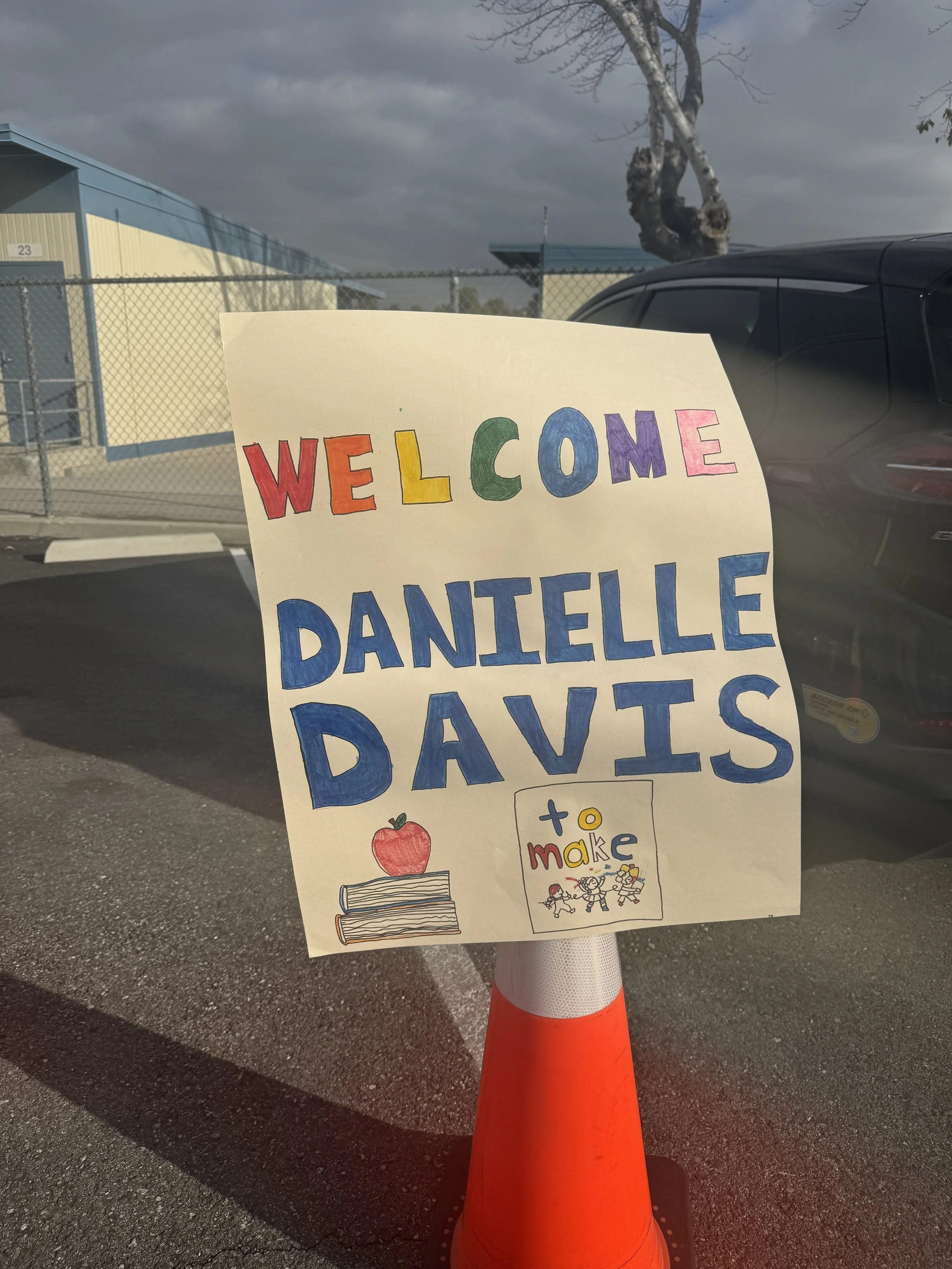 Colorful welcome sign on a traffic cone, featuring illustrations of a red apple, books, and a children’s drawing with the text 'Welcome Danielle Parents' for a school or community event.