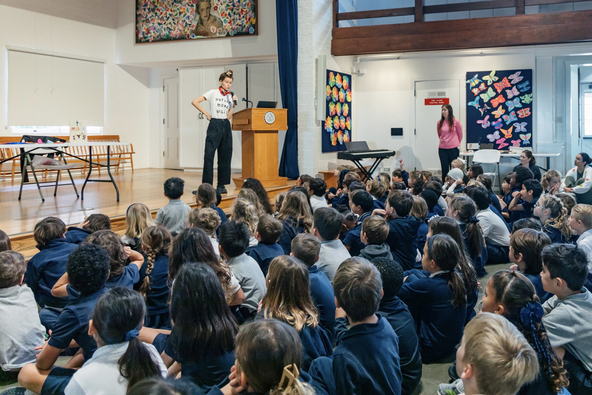 A woman standing on a stage near a podium, speaking into a microphone in front of a large group of children seated on the floor, with colorful artwork hanging on the walls.