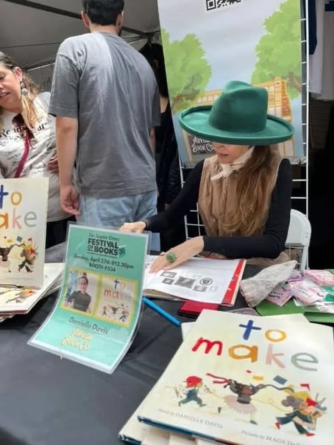 A woman wearing a large green hat sitting at a table with signs and books, at a festival booth with people and trees in the background.