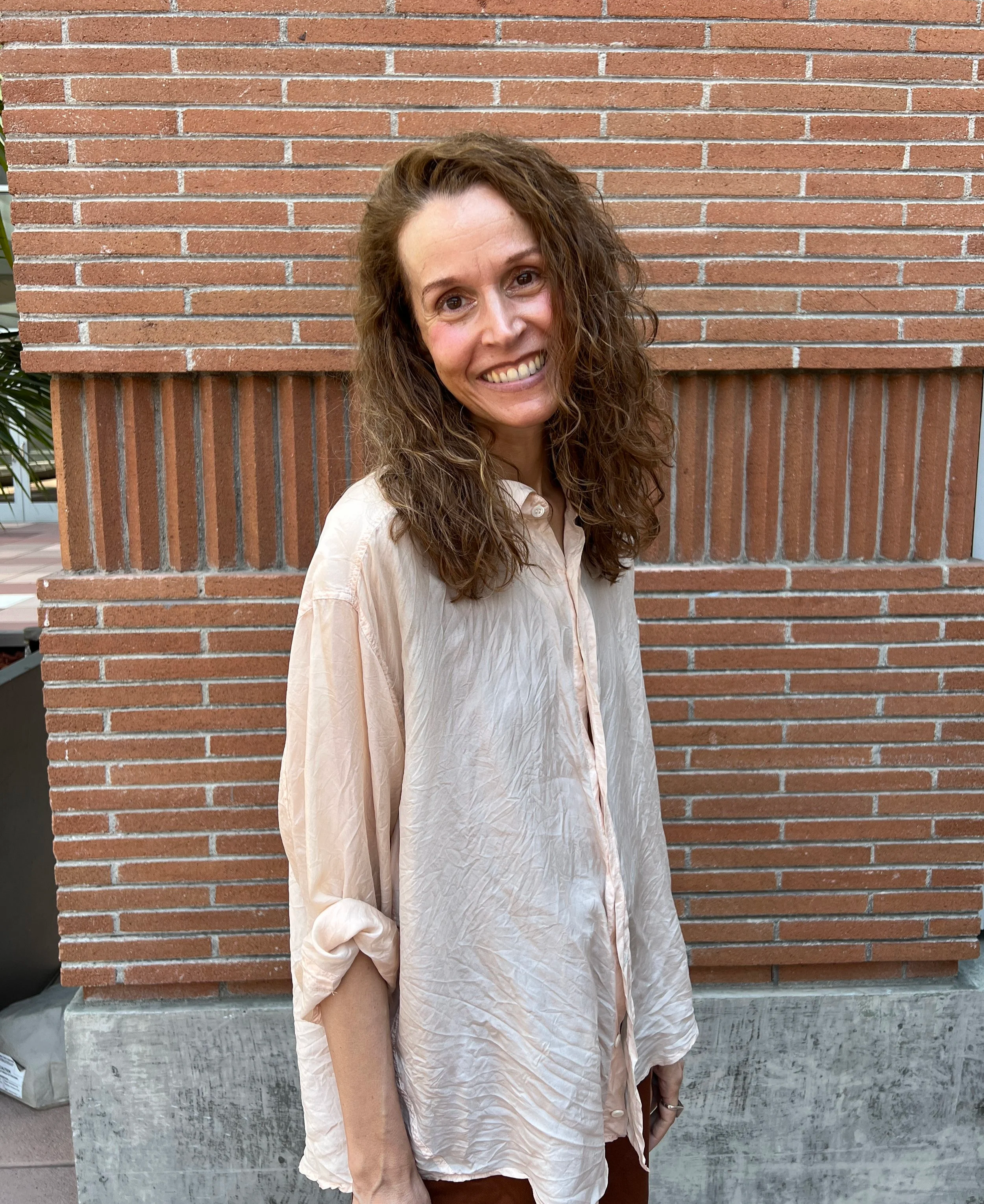 A woman with curly brown hair wearing a loose, cream-colored shirt standing in front of a brick wall.