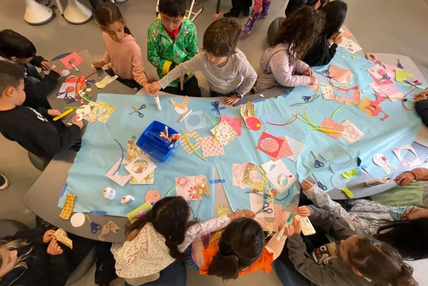 Children engaged in a craft activity at a large table covered with blue paper, various colorful paper cut-outs, scissors, glue, and ribbon.