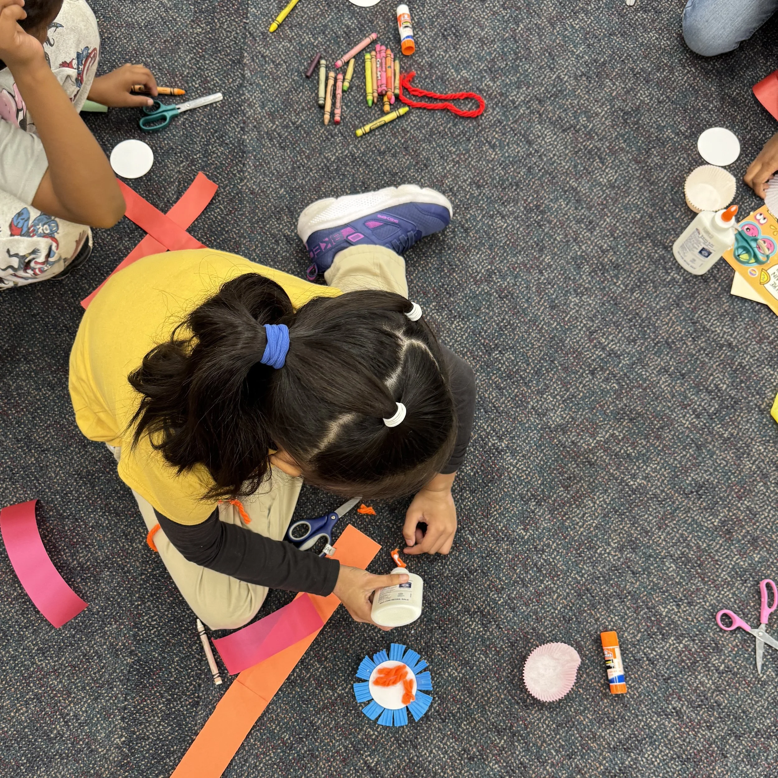 Children are sitting on the floor and creating crafts with paper, glue, scissors, and other craft supplies. Decorations and tools are scattered around, including a rolled strip of pink paper, cupcake liners, and paper cutouts.