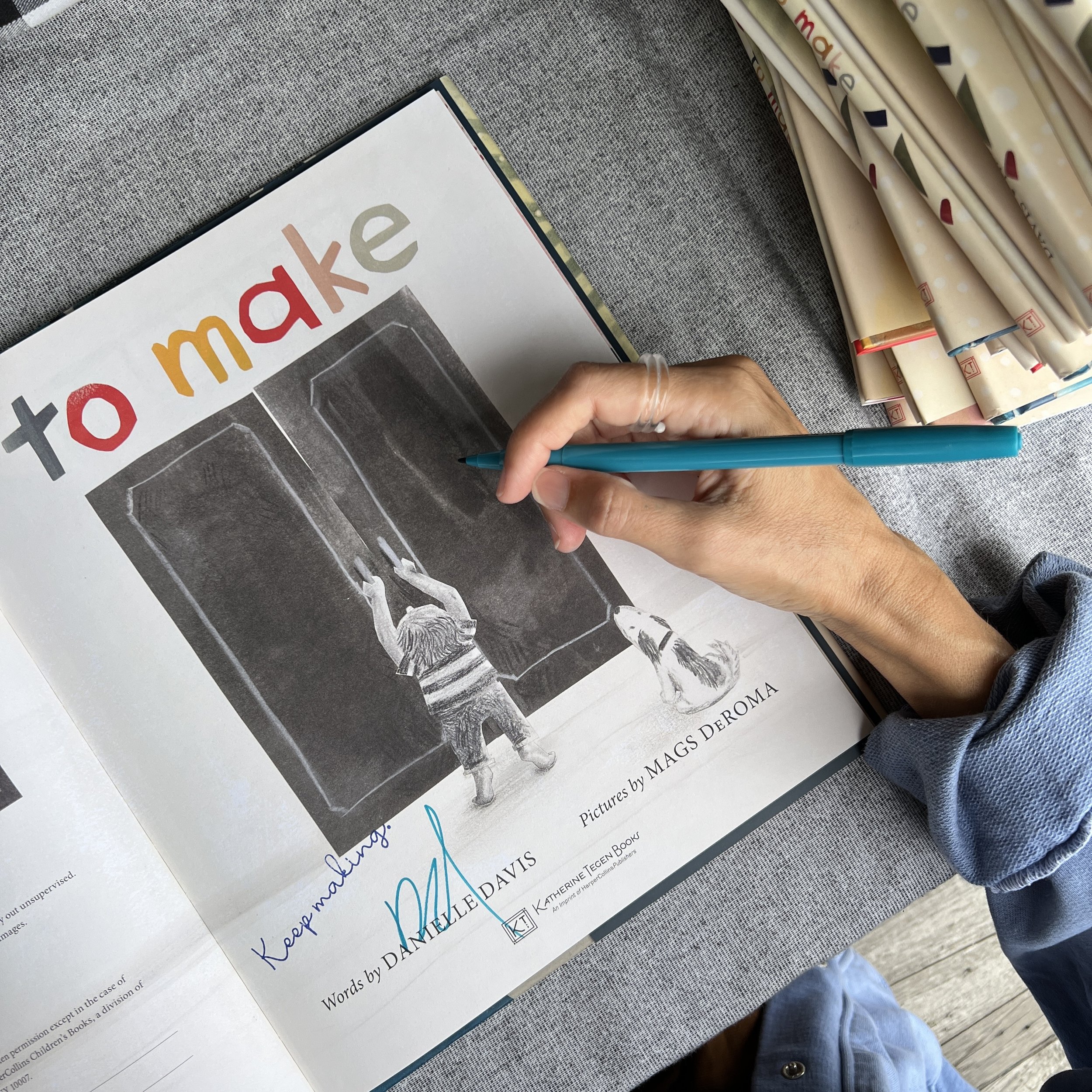 Close-up of a person holding a turquoise pen, working on a page from a book or magazine, with a stack of rolled papers in the background and a gray fabric surface underneath.