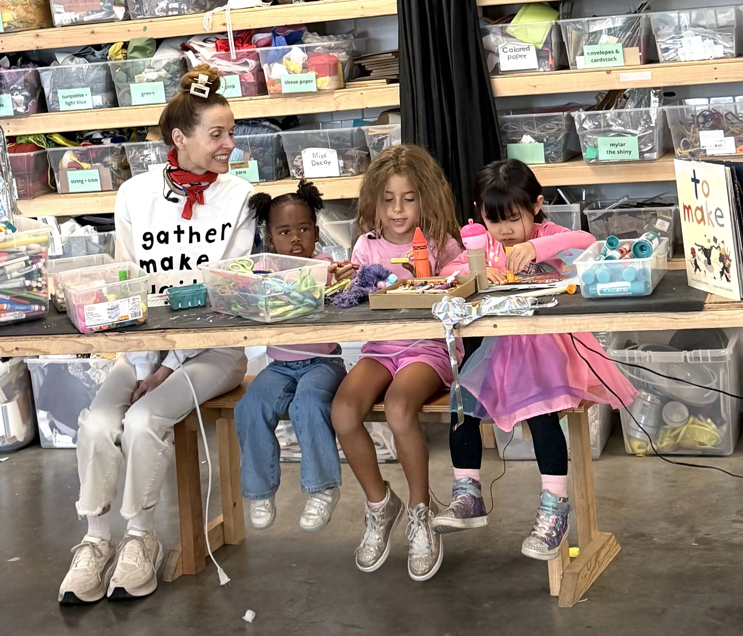 A group of four children sitting underneath a wooden table in a craft room, with a woman standing behind them. The shelves behind them are filled with labeled clear plastic bins containing various arts and crafts supplies.