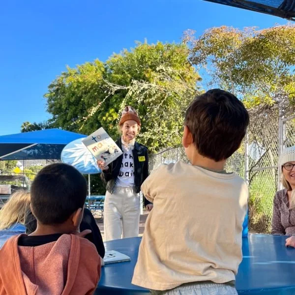 A woman reading a book to a group of children outdoors under a clear blue sky with trees in the background.