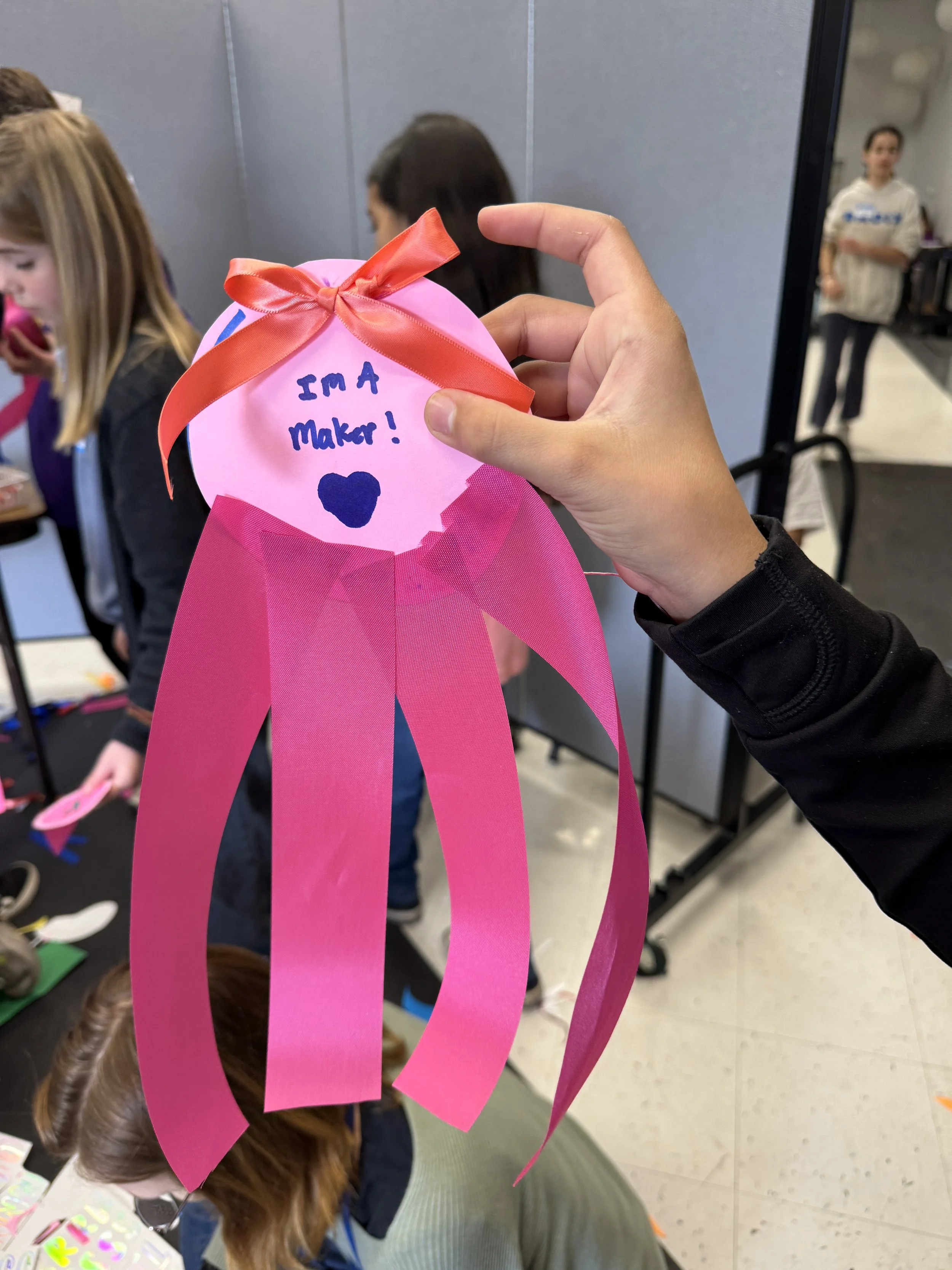 A pink paper ornament with a red ribbon bow, handwritten message reading 'I'm a Maker!' and a heart, held by a person's hand.