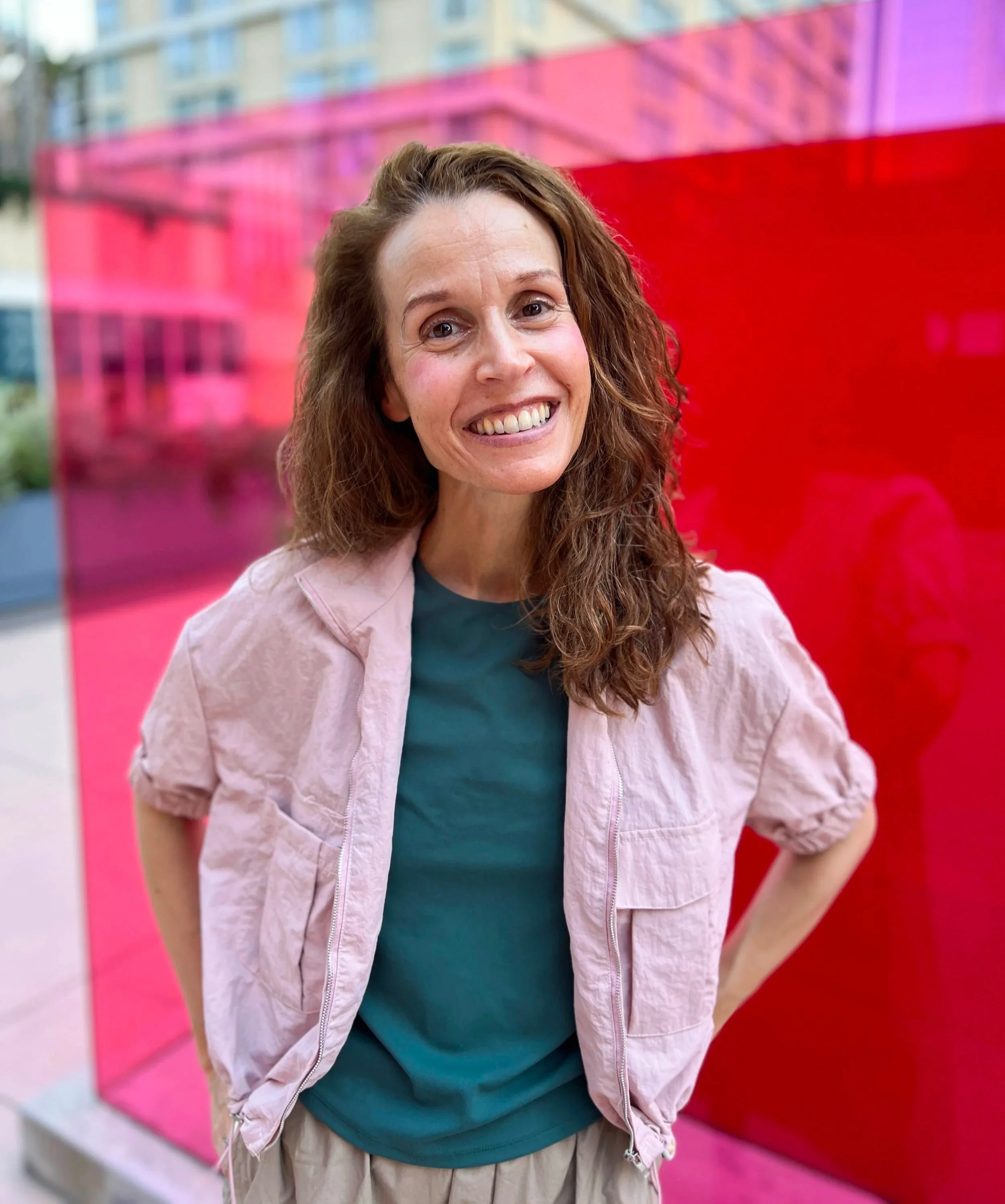 Smiling woman with long curly hair wearing a pink jacket and teal shirt standing in front of a pink and red wall.