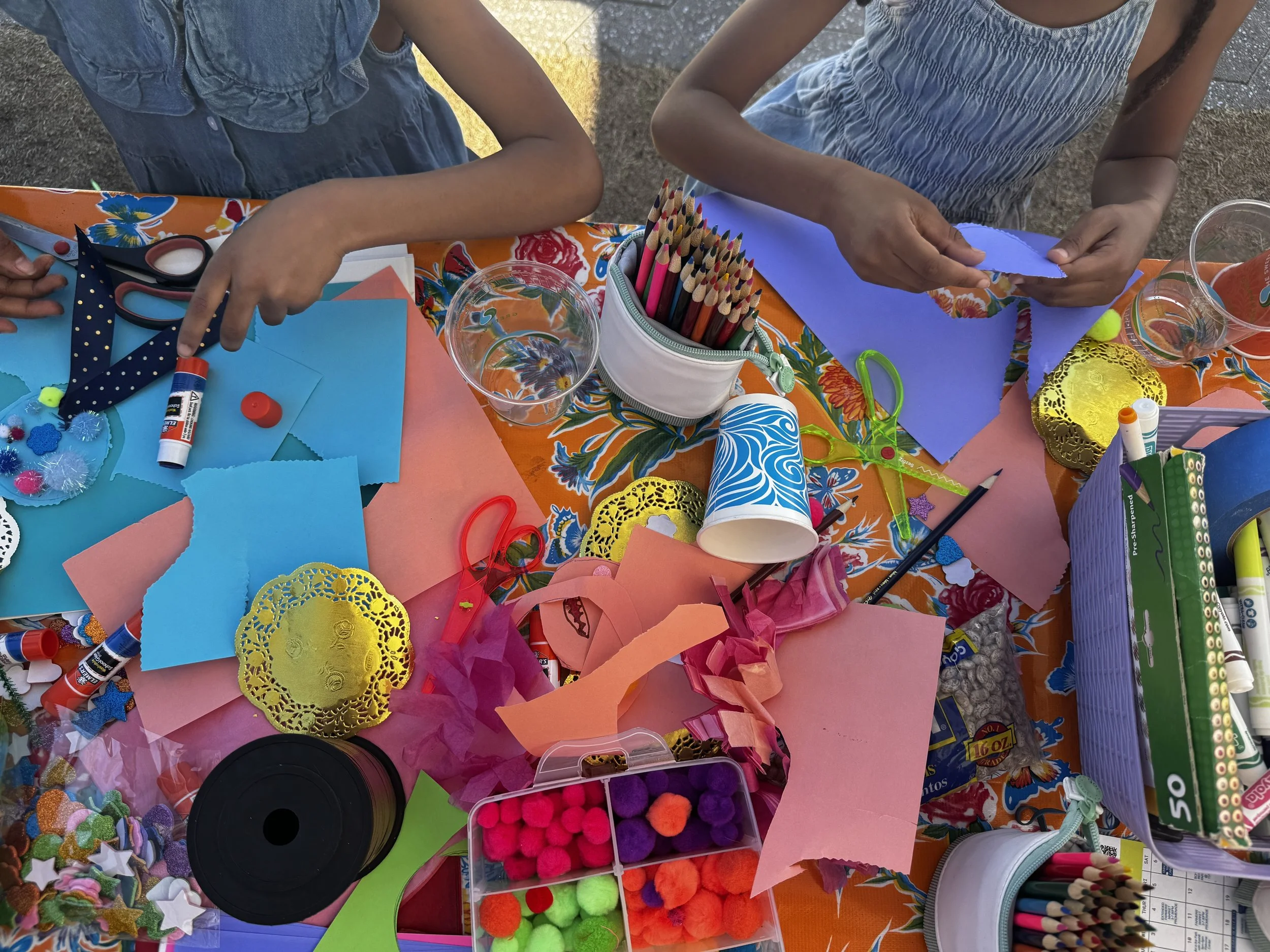 A table filled with colorful craft supplies, including colored paper, scissors, glue sticks, markers, pom-poms, and decorative items, set outdoors.