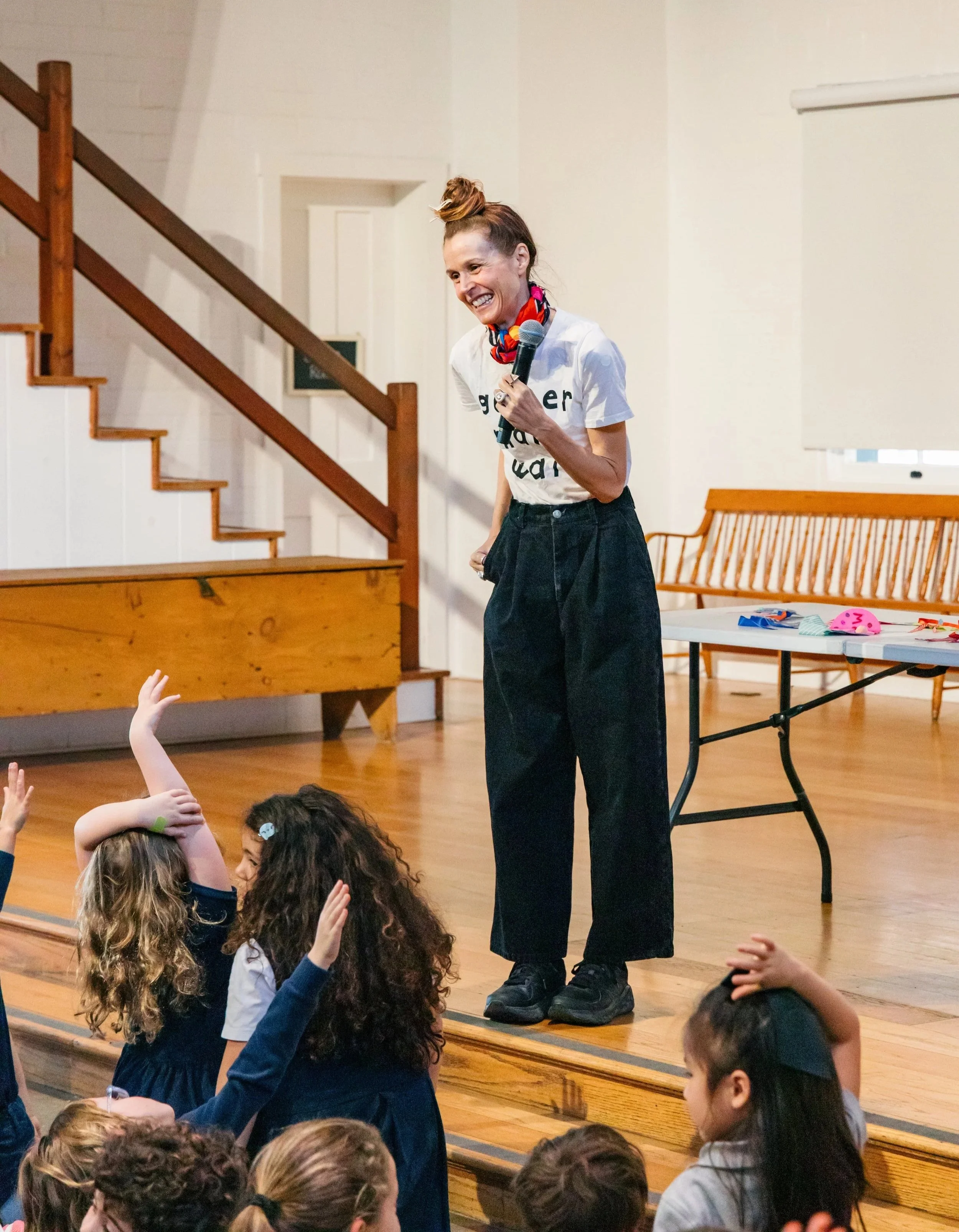 A woman with a bun hairstyle, wearing a white T-shirt and loose black pants, holding a microphone, smiling, and speaking to a group of children seated on the floor, raising their hands, in a room with wooden stairs and furniture.