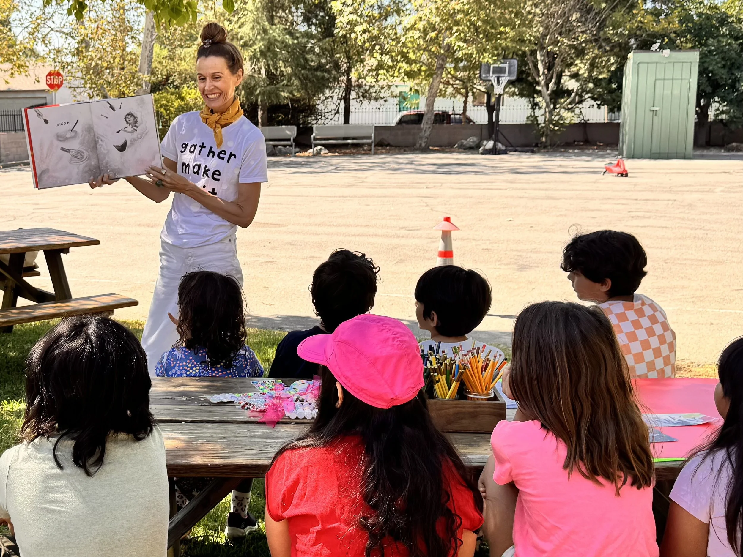 A woman standing outdoors in front of a group of children, holding and reading a picture book. The children sit on the ground, facing her, with some wearing colorful hats. There is a picnic table with art supplies nearby. In the background, there are trees, a basketball hoop, and a small green storage shed.