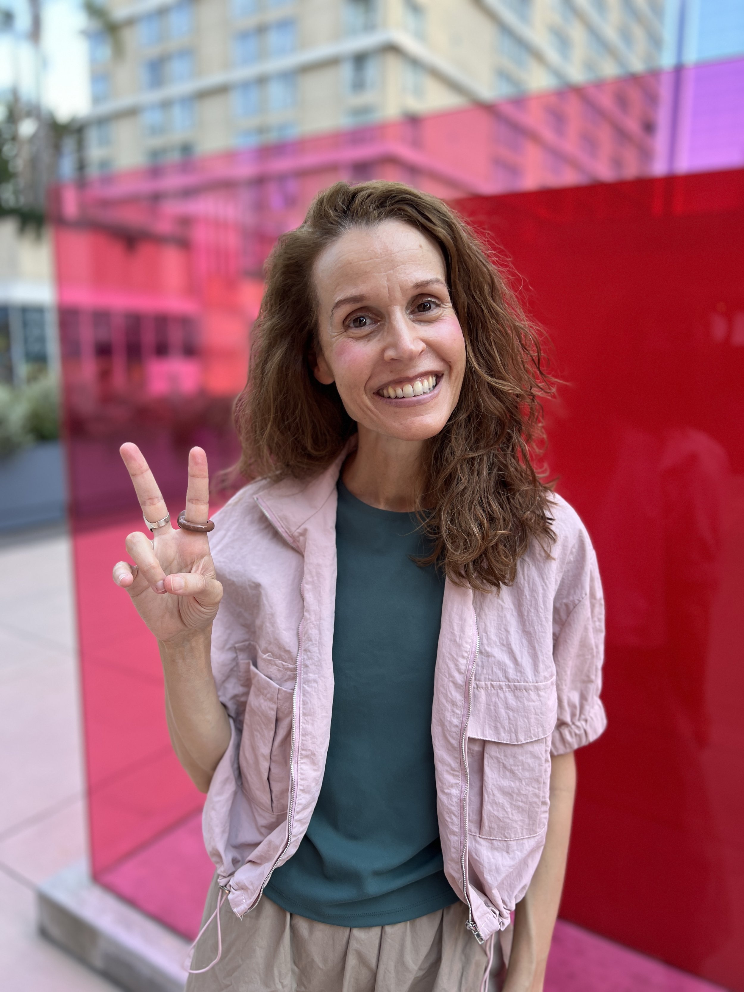 Danielle with wavy brown hair and a big smile showing her teeth, making a peace sign with her fingers, standing outdoors near a colorful pink and purple installation or sculpture, with city buildings in the background.