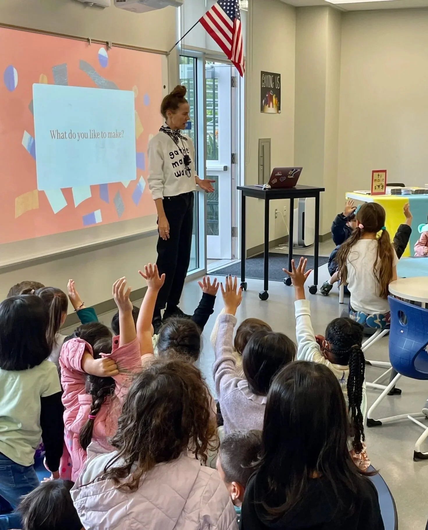 A woman leading a classroom activity with children sitting on the floor and raising their hands. A large screen behind her displays the question, 'What do you like to make?' There is an American flag mounted on the wall, and the classroom has a yellow table with stacks of papers or books.
