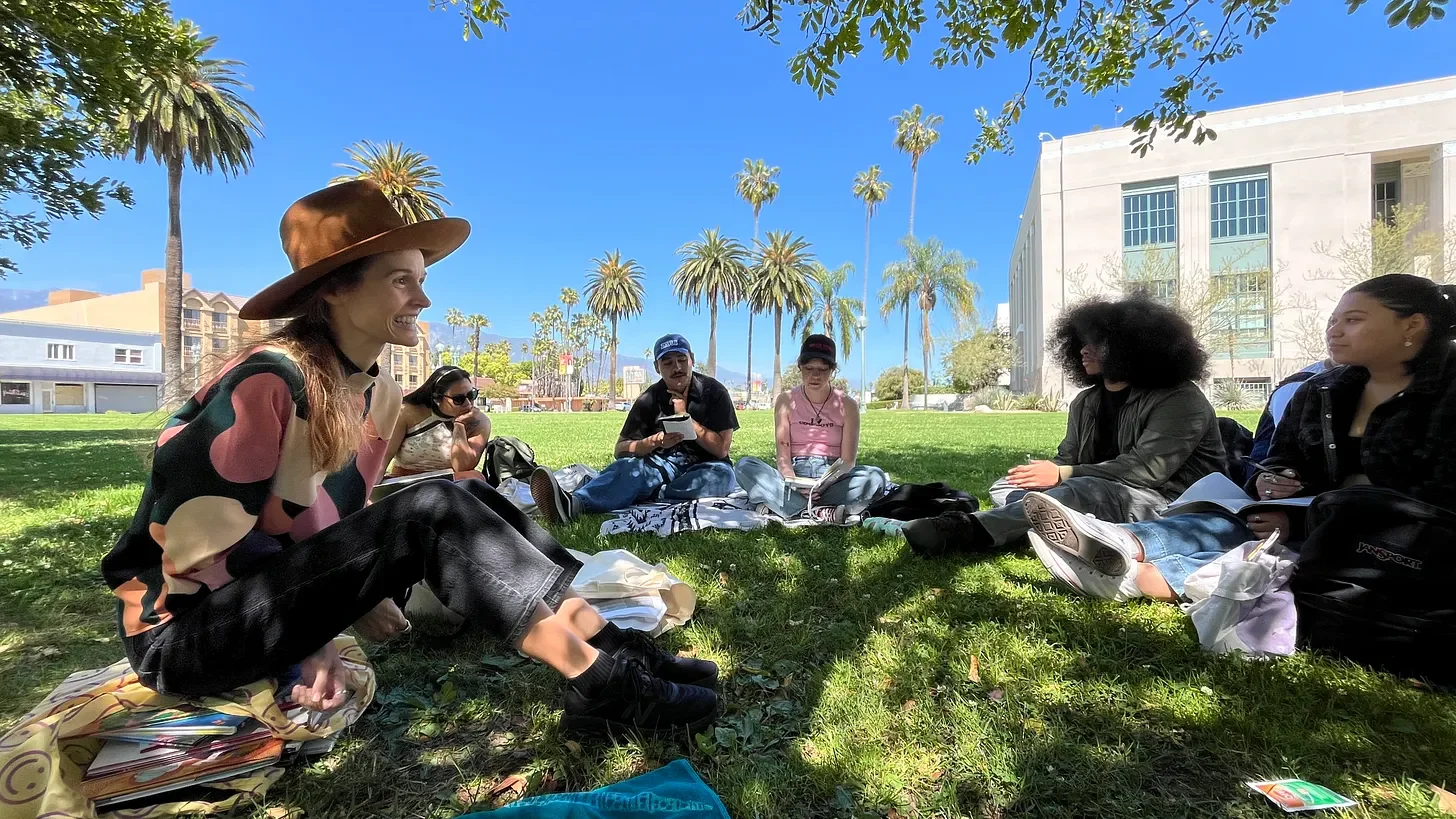 Group of people sitting cross-legged on a grassy park under trees, having a conversation on a sunny day with palm trees and a white building in the background.