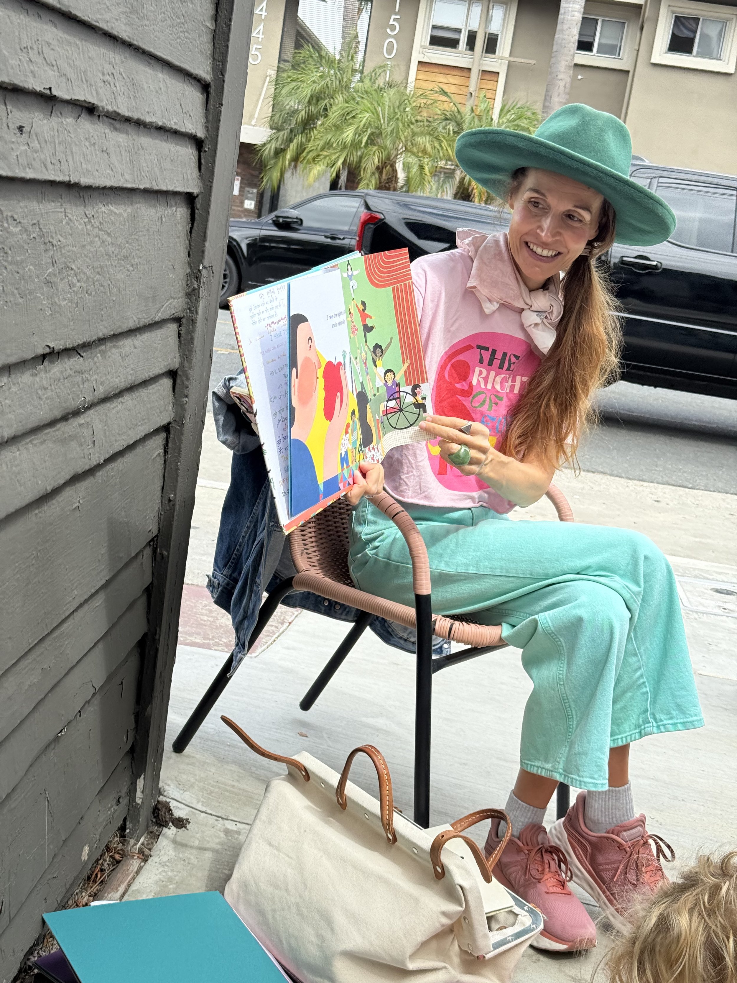 A woman with long reddish hair wearing a large green hat, pink shirt with the words 'The Right of' and mint green pants, sitting on a patio holding an open children's book and smiling. There are bags, a blue folder, and children partially visible nearby.