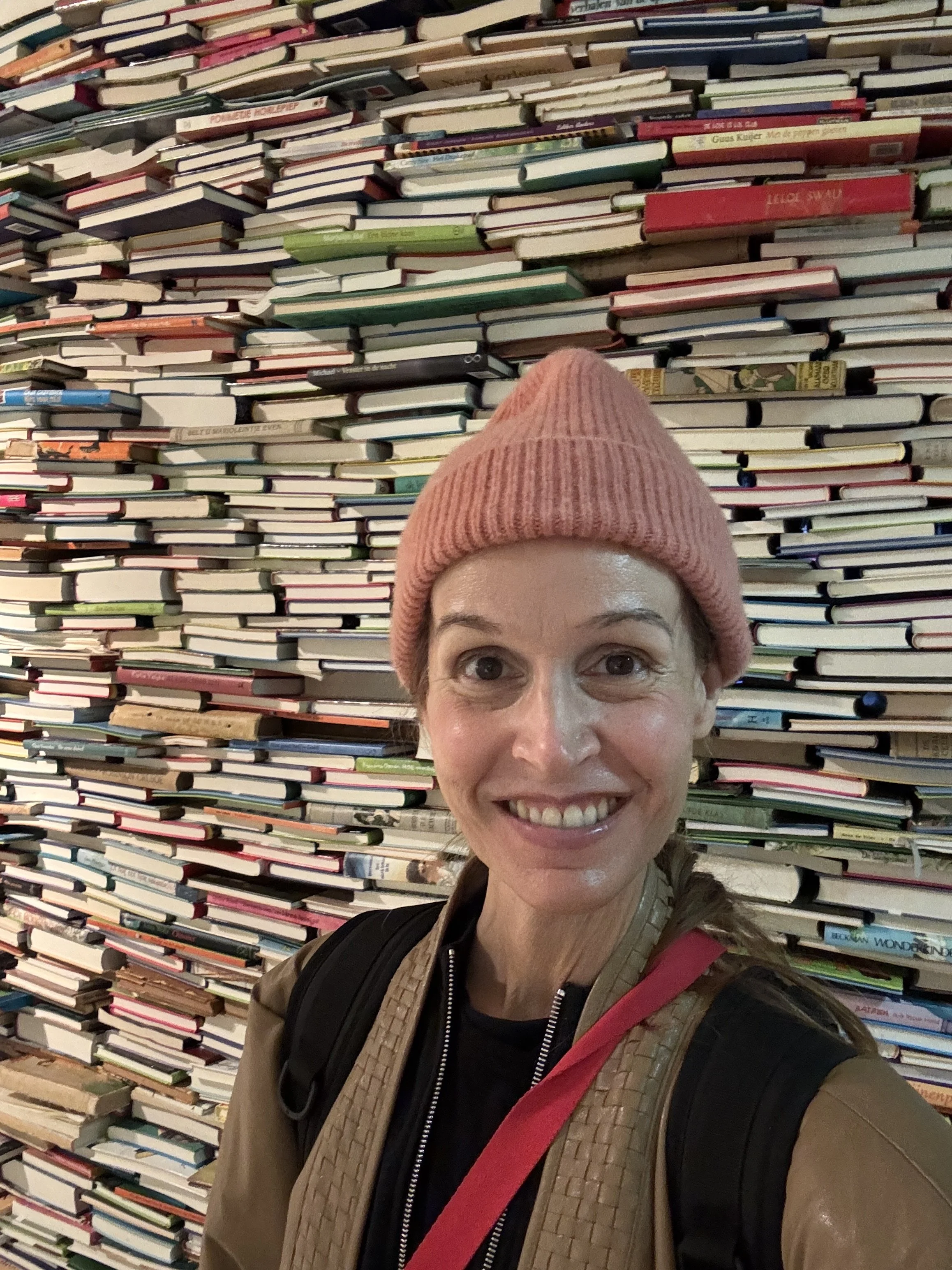 A woman wearing a pink knit hat and a brown jacket taking a selfie in front of a large wall of stacked books in a bookstore.