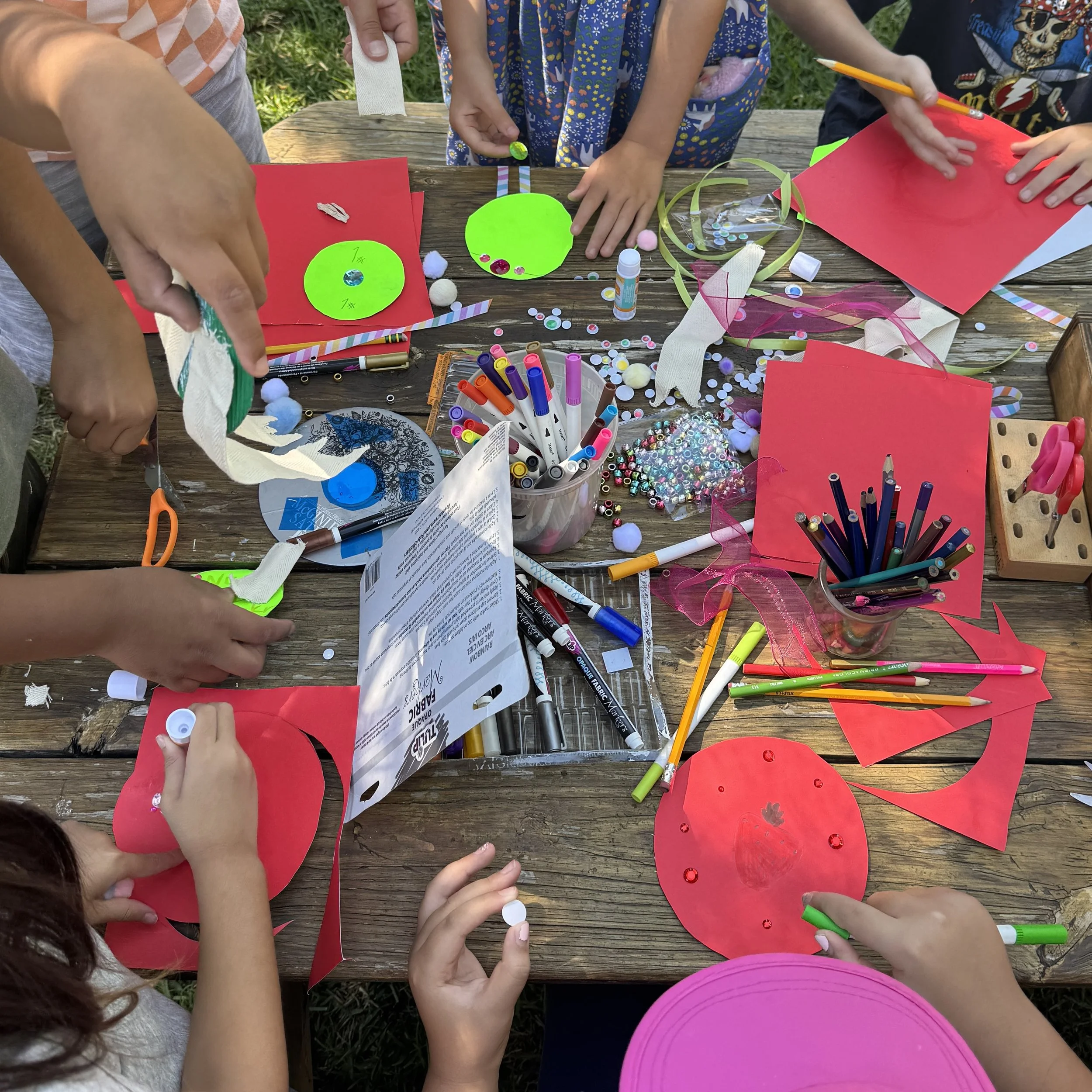 Children engaged in arts and crafts activities outdoors at a wooden table. They are using various art supplies such as colored markers, scissors, tape, and decorative materials like sequins, pom-poms, and ribbons. The setting appears to be a grassy outdoor area.