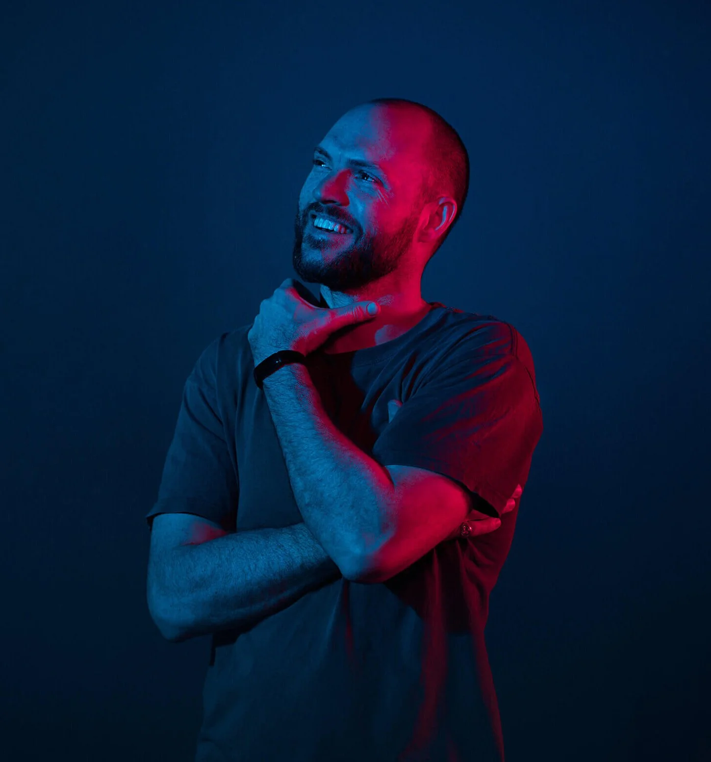 A smiling bald man with a beard, wearing a gray t-shirt and a black wristband, is posing with one hand on his neck and the other crossed over his chest against a dark background with red and blue lighting.