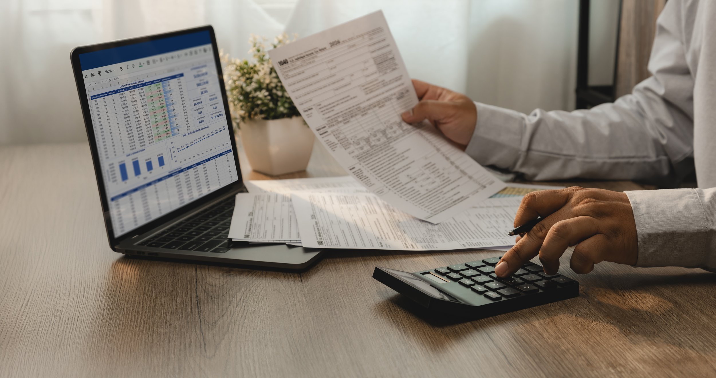 Persona revisando documentos y usando calculadora con una laptop que muestra gráficos y datos financieros.