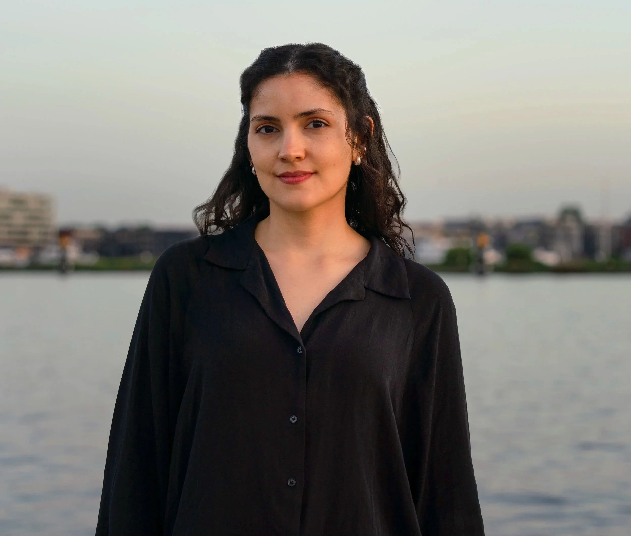 A woman with dark, wavy hair and earrings standing outdoors near a body of water with an urban skyline in the background.