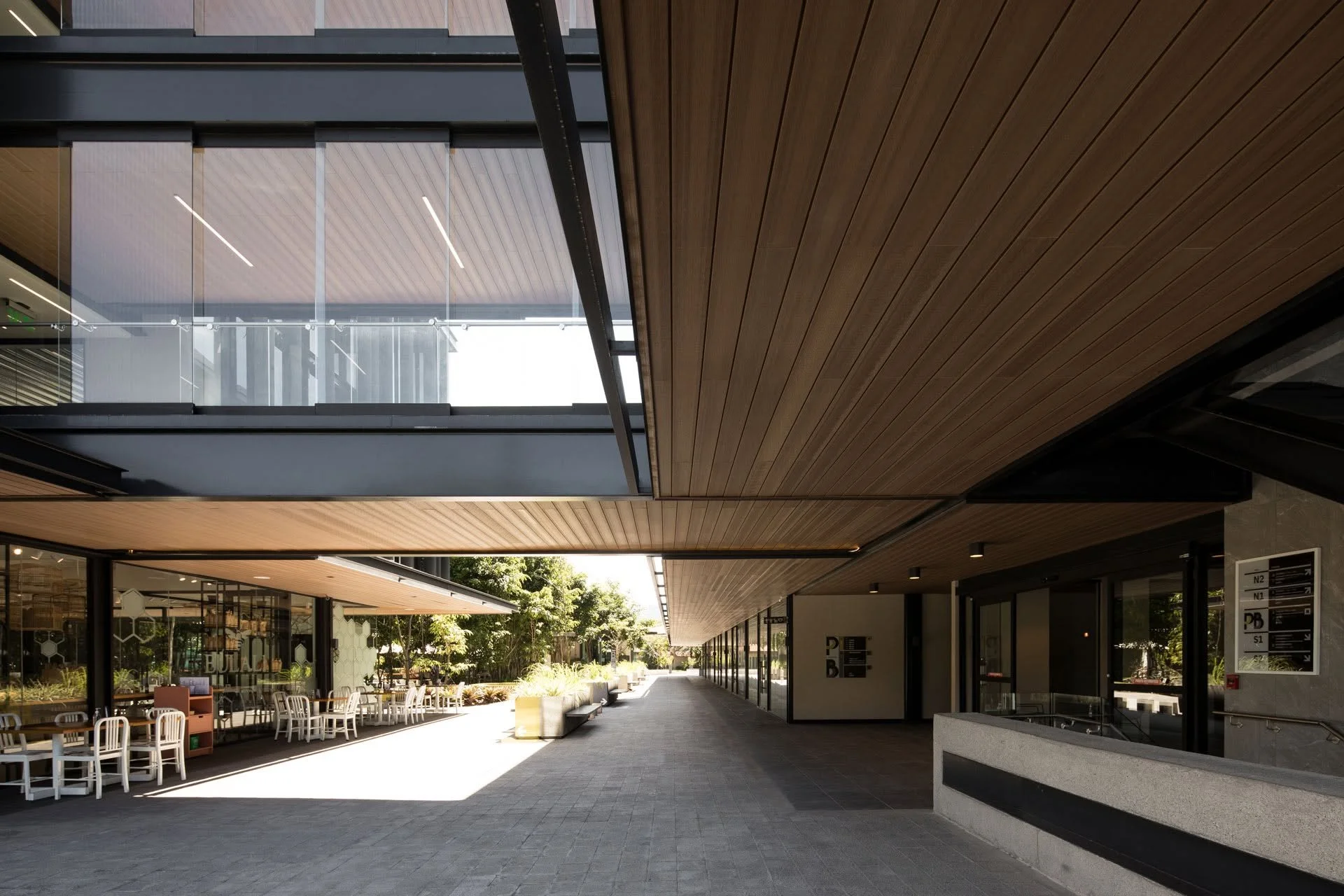 Pasillo cubierto de un complejo moderno con techos de madera, fachadas de vidrio, cafetería al aire libre, vegetación y señalización direccional.