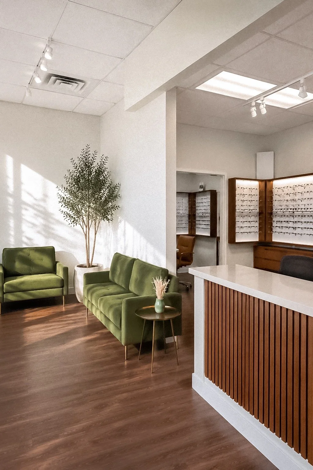 A waiting area with green sofas, a potted plant, and a wooden counter inside an office or clinic with eye prescriptions displayed on wall shelves in the background.