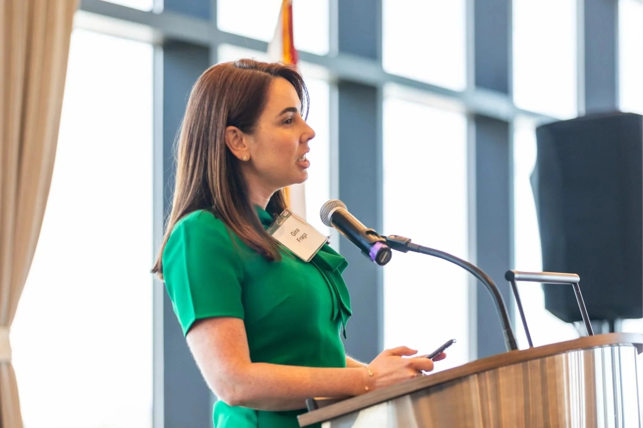 Attorney Gina Fraga with brown hair in a green dress speaking at a podium with a microphone, holding a smartphone, in a room with large windows.